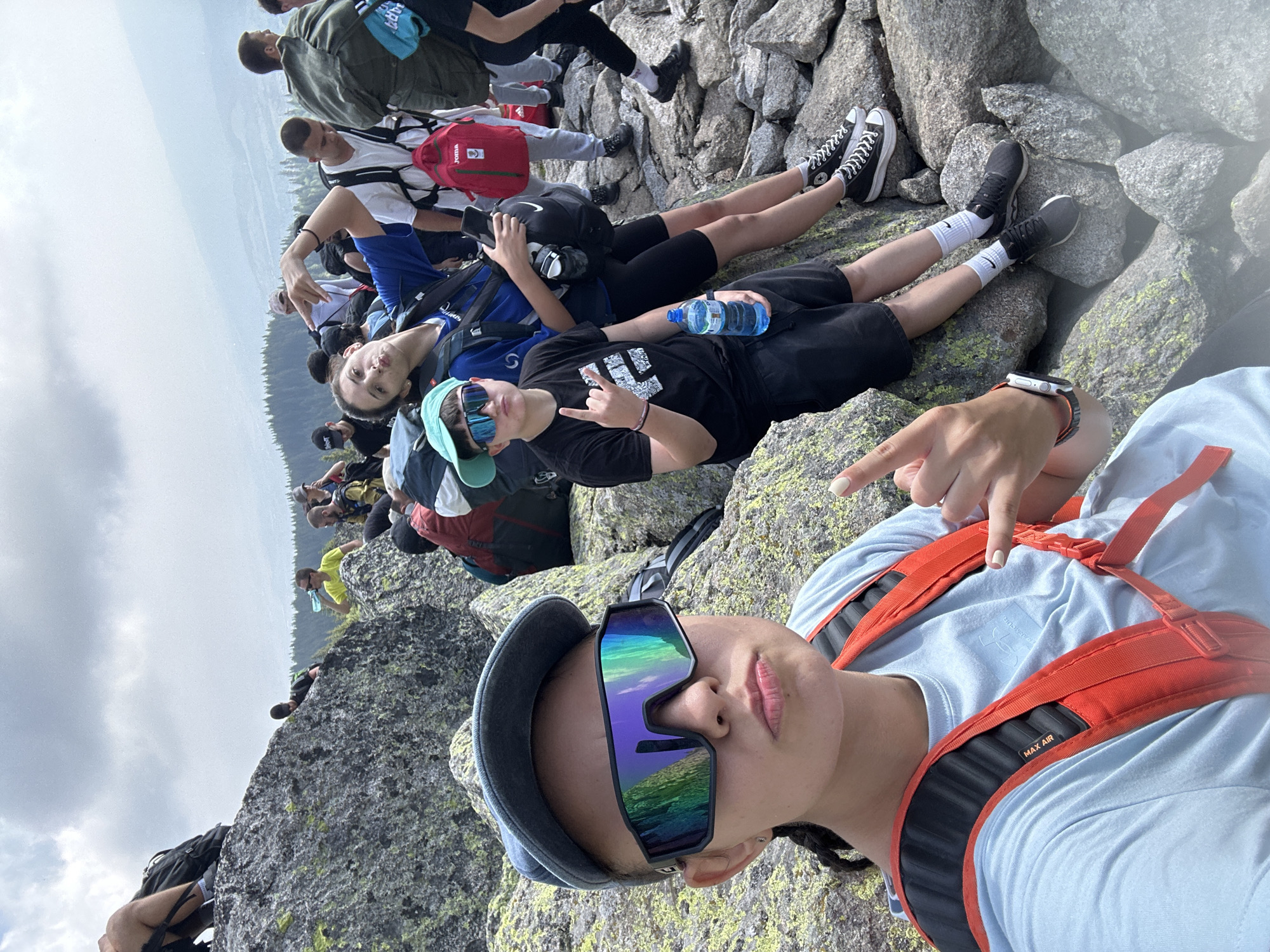The image depicts a group of young people on a rocky, mountainous terrain, likely in the vicinity of Bistritsa, Bulgaria, given the backdrop of forested hills and a distant, hazy mountain range. In the foreground, a person wearing a light blue t-shirt, a grey baseball cap, and reflective, oversized sunglasses is taking a selfie. They are also wearing a bright orange backpack with black straps. Their left hand is making a rock-on gesture, while their right hand is partially visible, also making a similar gesture. To the right of the selfie-taker, another young person in a blue t-shirt and a turquoise cap is seated, holding a smartphone and looking towards the camera. A third person, wearing a black t-shirt and shorts, is also seated to their left, holding a bottle of water. This individual is giving a peace sign with their left hand and pointing with their right. Several other individuals are visible in the background, some carrying backpacks, sitting on rocks, or standing. The environment is characterized by large, grey boulders, sparse vegetation, and a cloudy sky. The overall scene suggests a casual outdoor gathering, possibly a hike or a rest stop during an excursion.