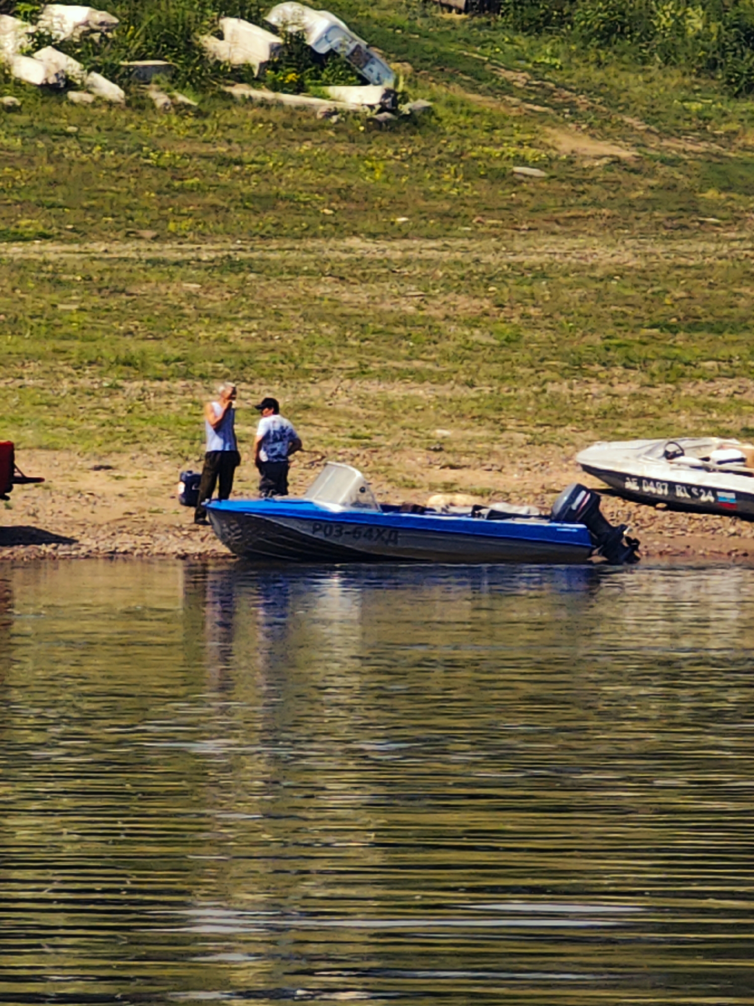 Two men stand on a sandy bank next to a blue motorboat docked at the water's edge. The man on the left wears a light blue sleeveless shirt and dark pants, holding a dark object to his ear, possibly a phone. The man on the right wears a dark shirt with a white pattern and dark pants, and has a baseball cap on his head. The blue motorboat has the registration number "P03-64XH" visible on its side. Further to the right, a second motorboat, white with a dark stripe and registration "NE 0497 RL$24", is also beached. The background consists of a grassy slope with scattered white debris and vegetation. The water in the foreground is rippled and reflects the surrounding environment. The scene appears to be outdoors, likely near a body of water, given the presence of boats and the shoreline. The location of Погодаево, Russia, is not discernible from the visual elements within the image itself.