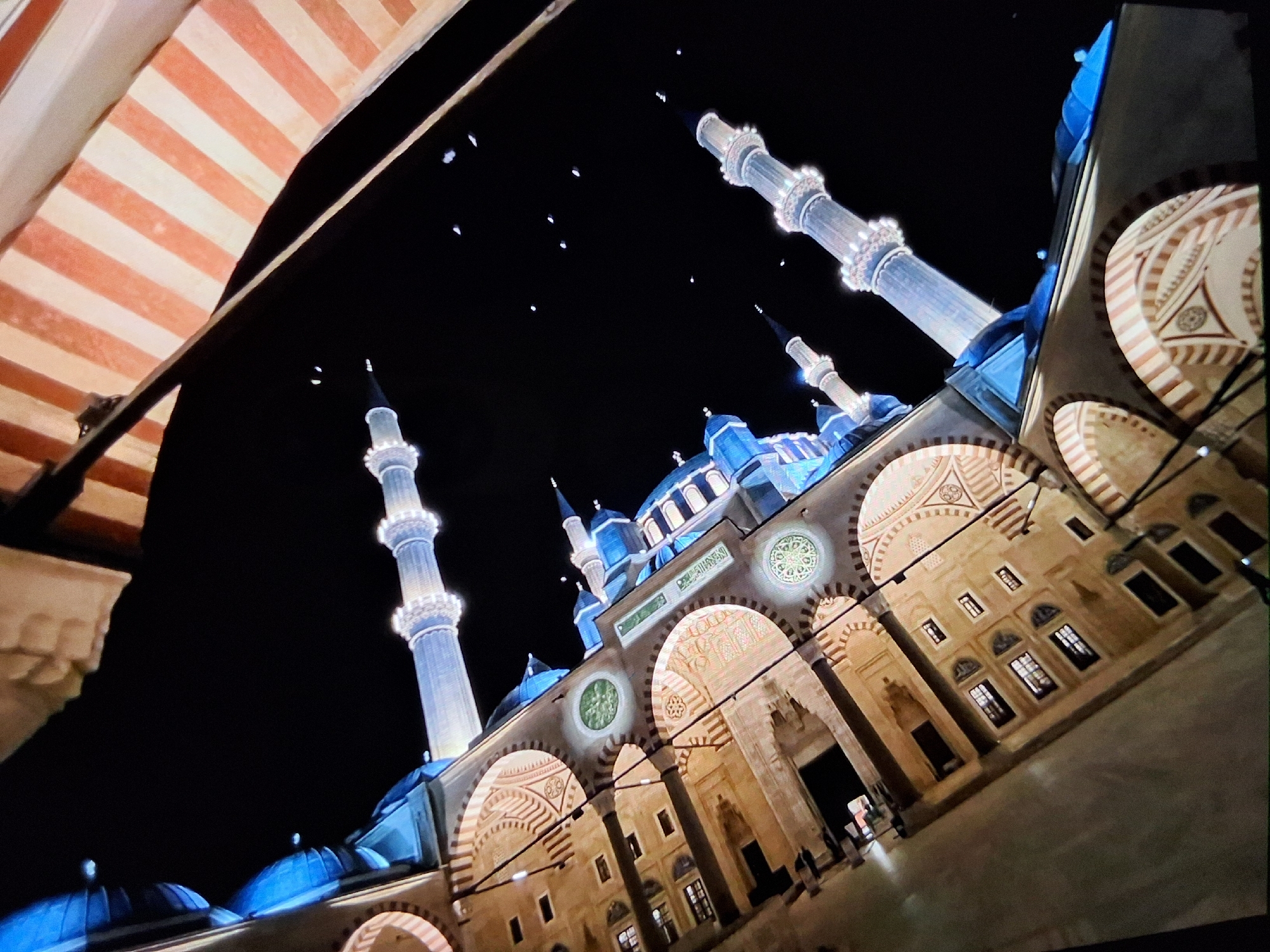 An angled, low-light photograph captures the illuminated minarets and facade of a mosque at night, against a dark sky dotted with a few small, bright specks. The architectural details of the mosque, including arched windows and ornate decorative patterns, are highlighted by warm, golden light, while the domed sections and minarets are bathed in cool blue light. In the upper right corner, a portion of an awning with red and white stripes is visible, creating a diagonal division in the frame. The overall composition is skewed, giving a dynamic perspective of the building.