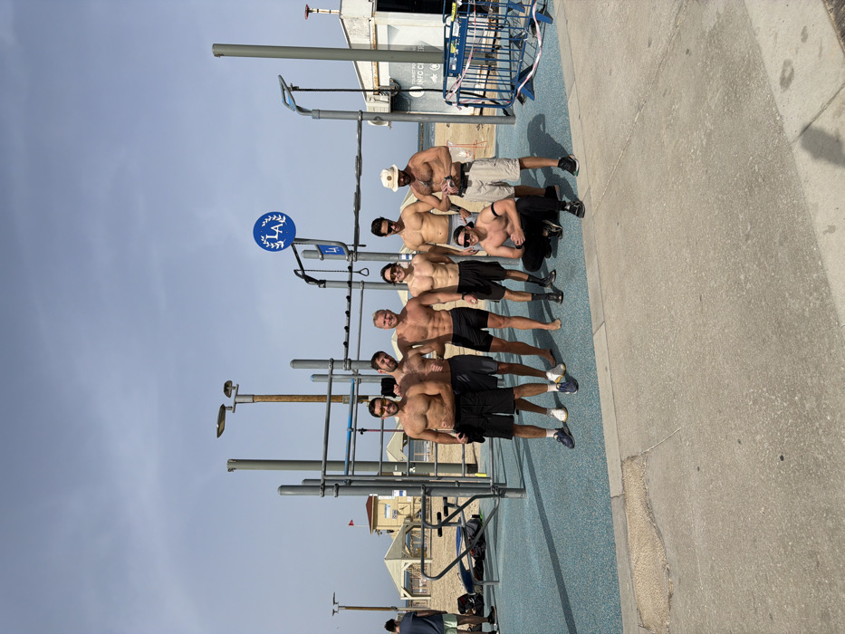 An outdoor scene captures eight adult males posing for a photograph at a beachside calisthenics park in Tel-Aviv, Israel. Seven men stand, while one crouches in the immediate foreground. All individuals are shirtless, revealing muscular physiques, and wear athletic shorts or swim trunks; most have athletic shoes, with one individual standing barefoot.
The calisthenics park features a multi-station metal structure with various horizontal bars, parallel bars, and rings, set upon a blue, possibly rubberized, safety mat. A prominent blue circular sign, bearing a white laurel wreath and stylized "LA" (or "IA"), is affixed to a pole within the gym apparatus.
Behind the gym, a sandy beach extends towards the sea, visible in the background. Several white-roofed shaded structures and a distinct hexagonal building with a red flag pole, likely a lifeguard station, are situated on the sand further down the beach. To the right of the calisthenics park, a light-colored building, possibly a kiosk or information center, displays a partially obscured sign reading "TOURIST INFO." Blue temporary barriers and red-and-white tape are positioned near this building. A concrete pathway occupies the immediate foreground. The sky is partly cloudy, suggesting daytime conditions.