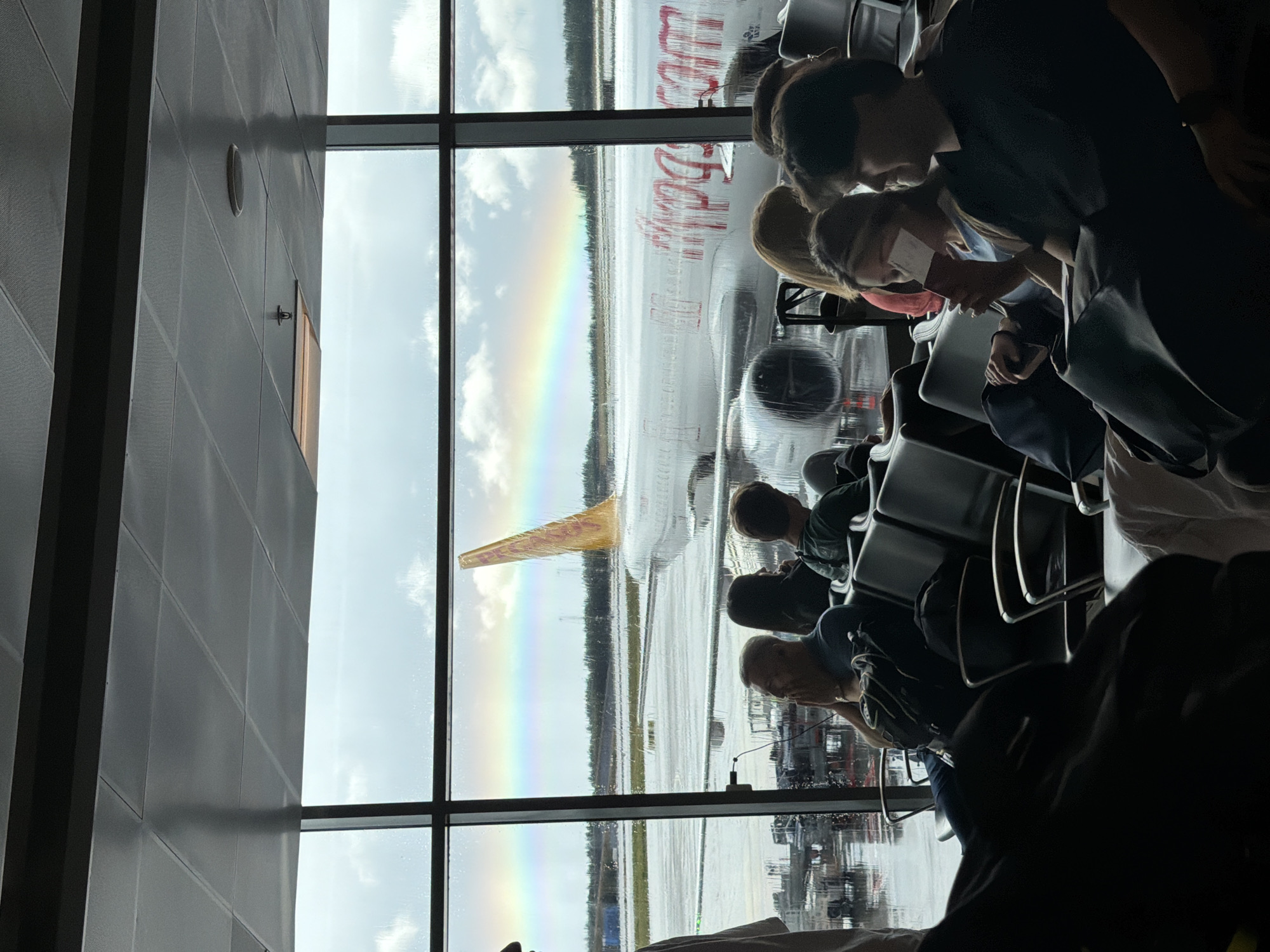 The image depicts the interior of an airport terminal in Gardermoen, Norway, specifically a departure lounge. Large windows offer a view of the tarmac, where an airplane is parked. The weather outside appears to be rainy, with water streaks visible on the window panes and a rainbow arching across the sky.
Inside the terminal, several passengers are seated on dark, upholstered chairs, waiting for their flights. The lighting is dim, with the primary illumination coming from the overcast sky outside and some overhead fixtures.
In the foreground, the backs of several passengers are visible, some with luggage. To the left, a person is standing, partially visible. In the middle ground, passengers are seated, some looking out the window, others engaged with their devices or simply waiting. One woman is holding a passport.
The airplane outside is predominantly white with red lettering that appears to spell out "pegasus.com." The tail fin of the plane is a bright yellow and orange. The tarmac is wet, reflecting the subdued light. In the background, other airport infrastructure and buildings can be seen. The ceiling of the terminal is visible at the top of the image, composed of acoustic tiles.