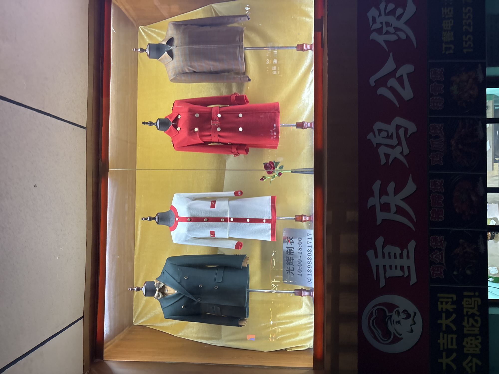 This image is a medium shot of a storefront display in the Yuzhong District of China. The display features four mannequins dressed in formal attire. The first mannequin on the left is wearing a dark green double-breasted suit with a tan shirt and a bow tie. The second mannequin is dressed in a white and red tweed suit with a skirt. The third mannequin is wearing a red trench coat with a belt and white buttons. The fourth mannequin, on the far right, is wearing a brown plaid jacket. Behind the mannequins, a golden fabric drapes the wall. A small sign with Chinese characters is visible below the first two mannequins, indicating business hours and a phone number. Below the display, a red sign with white Chinese characters reads "重庆鸡公煲" which translates to "Chongqing Gong Bao Chicken." The sign also includes smaller images of food and text that appears to be a phone number. The overall scene suggests a clothing store with a restaurant below it.