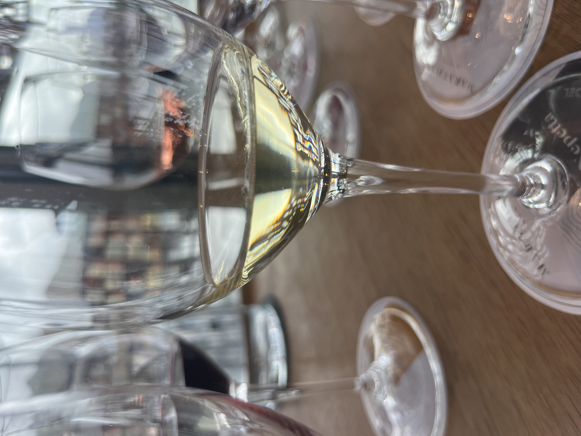 The image shows a close-up of wine glasses on a wooden table. The foreground is dominated by a clear wine glass filled with a pale yellow liquid, presumably white wine. The liquid reflects light, creating rainbow-like refractions on the glass. The stem of this glass is clear and appears to have some texture. Behind this glass, other wine glasses are visible, some containing red wine, and others appearing empty or containing a pinkish liquid. In the background, there is a blurred view of what appears to be an indoor setting with architectural elements and possibly distant scenery. The overall impression is that of a wine tasting or a meal at a restaurant or winery. The location is specified as Oikos, Cyprus.