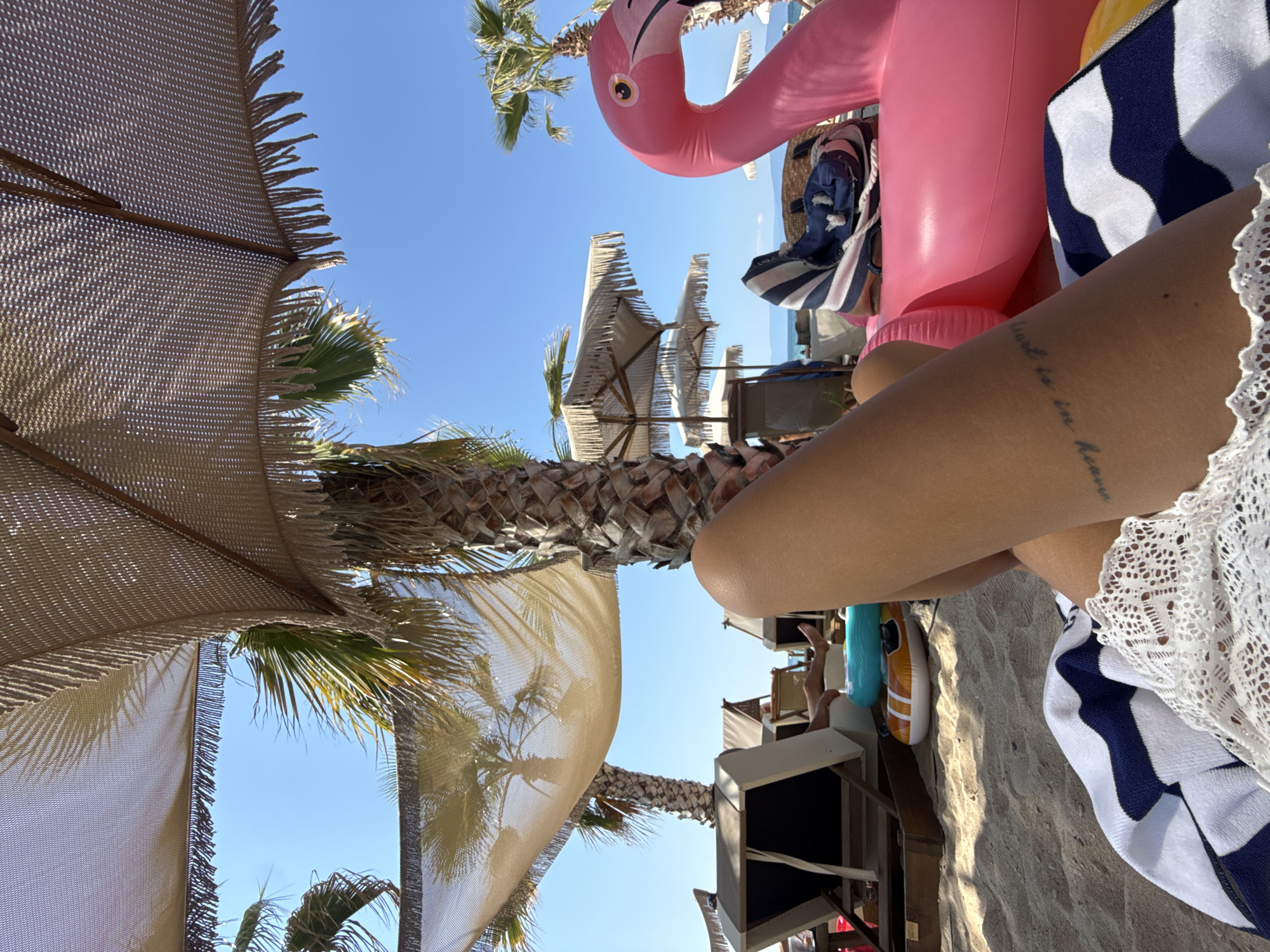 The image is taken at Ofrynio Beach, Greece, on a sunny day. The perspective is from a person lying down on the sand, looking upwards and outwards. In the foreground, the legs of the person are visible, with a tattoo on the left thigh. A striped blue and white towel and white lace garment are also in the foreground. To the right, a large, inflatable pink flamingo float is prominent. Beyond the float and the person, the sandy beach extends, with several beach chairs and umbrellas. Palm trees, some casting shadows from their fronds, are scattered throughout the scene. In the background, the blue sea and sky are visible, with a distant coastline under a clear blue sky. The overall impression is a relaxed beach scene.