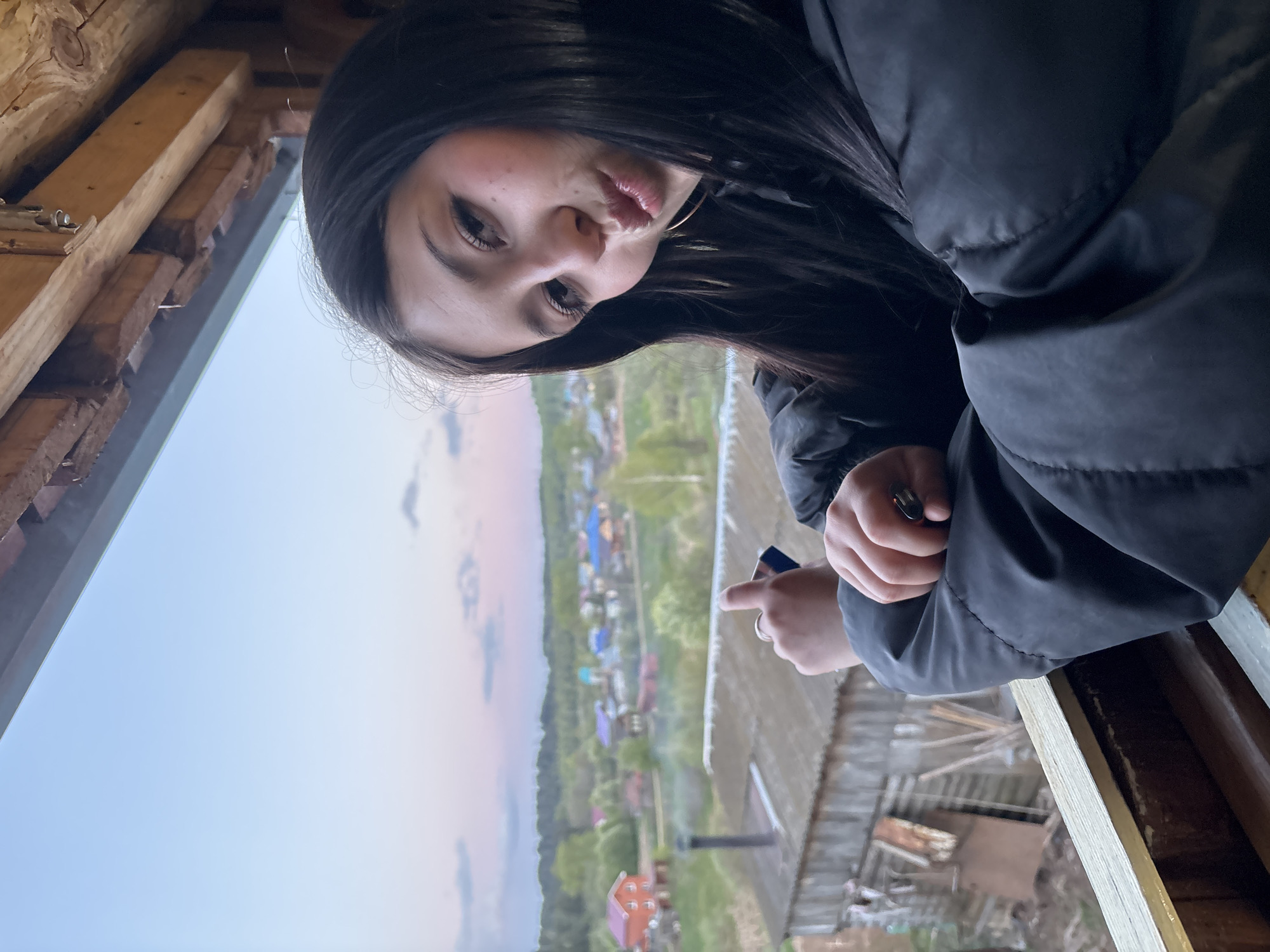 A young woman with long, dark hair is photographed leaning out of a window, possibly in a rural area. She is wearing a dark-colored, puffy jacket. Her lips are slightly pursed. Her right hand, adorned with a ring, holds a small dark object and her index finger is extended. Outside the window, a view of a village or small town stretches into the distance, featuring houses with colorful roofs interspersed with trees and greenery. The sky has a gradient of light blue and pink. The window is framed by unfinished wood, indicating the structure is likely under construction or renovation. Based on the context, the location might be in or near Фоки, Russia.