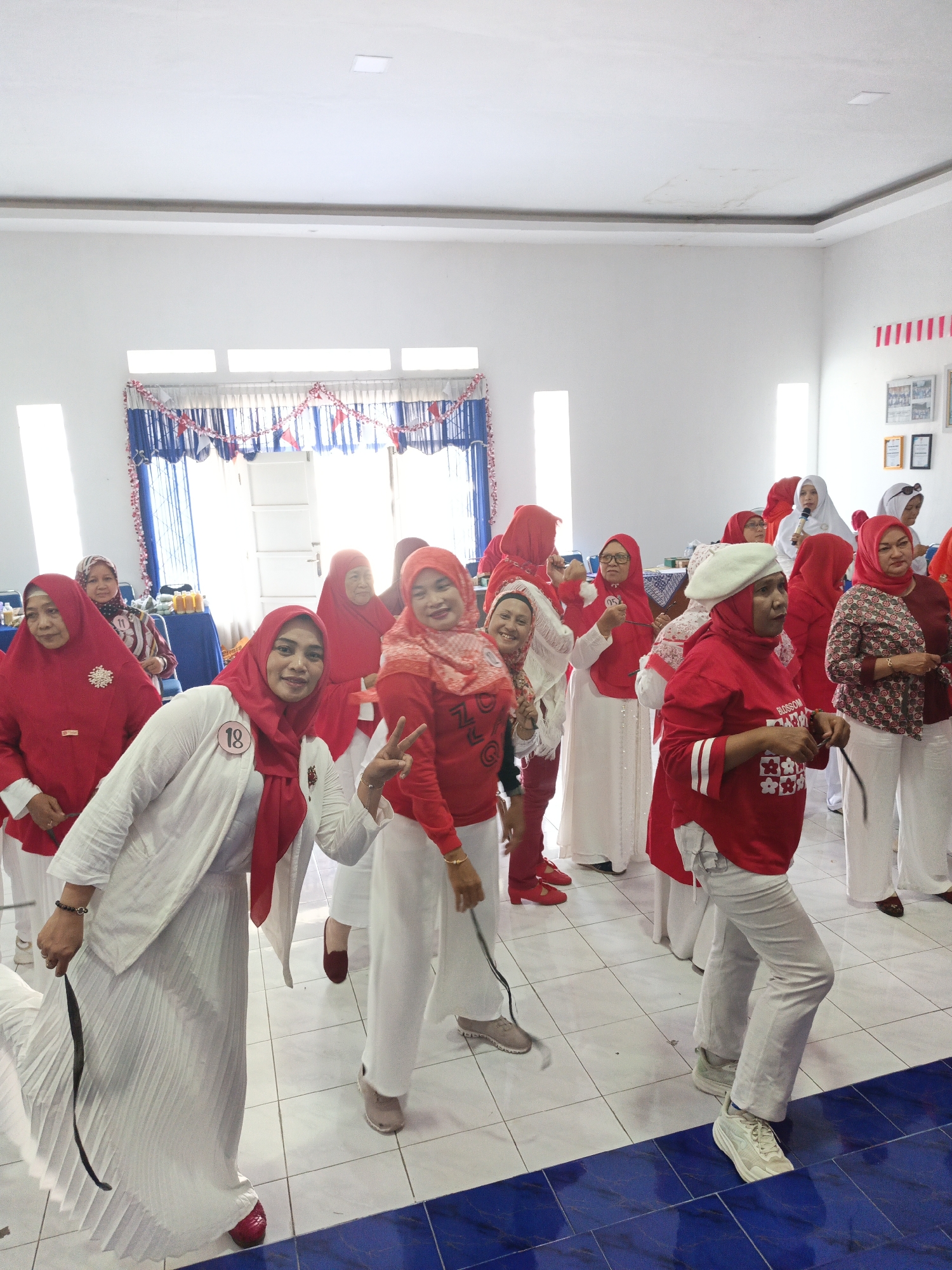 A group of women, most wearing red and white attire and headscarves, are gathered indoors, possibly for an event. The room has white walls and a tiled floor with blue accents. Decorations, including red and white flags, are visible in the background. Some women are smiling and posing for the camera, while others appear to be engaged in conversation or activity. One woman in the foreground is wearing a white jacket over a red headscarf and a white pleated skirt, holding a black ribbon. Another woman to her right is wearing a red shirt with white stripes and a white beret, also holding a black ribbon. The overall atmosphere suggests a festive or celebratory occasion. The presence of specific decorations and the attire of the participants hint at a possible Independence Day celebration in Indonesia. The location is specified as the city of Klaten, Indonesia.