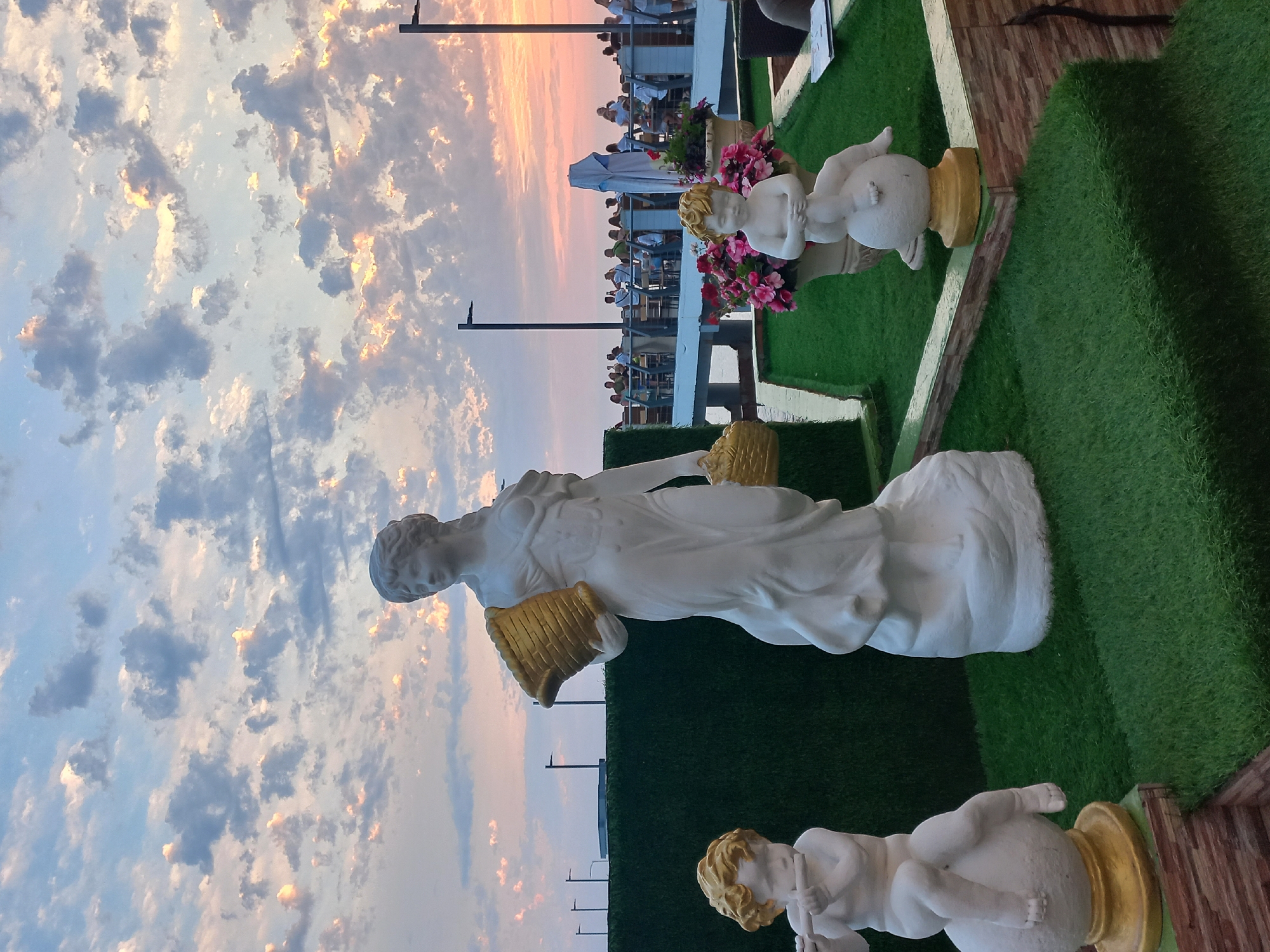 A rooftop restaurant in Sochi, Russia, is captured at sunset. The sky is filled with dramatic clouds painted in hues of pink, orange, and blue, indicating the sun is low on the horizon. In the foreground, white statues adorn the space, which is covered in artificial turf. To the left, a statue of a child plays a flute while sitting on a golden sphere. In the center, a larger statue of a woman holds a golden basket. To the right, another child statue sits on a golden sphere in front of flower pots filled with pink flowers. In the background, diners are seated at tables on a raised deck, with dark lamp posts interspersed along the railing.