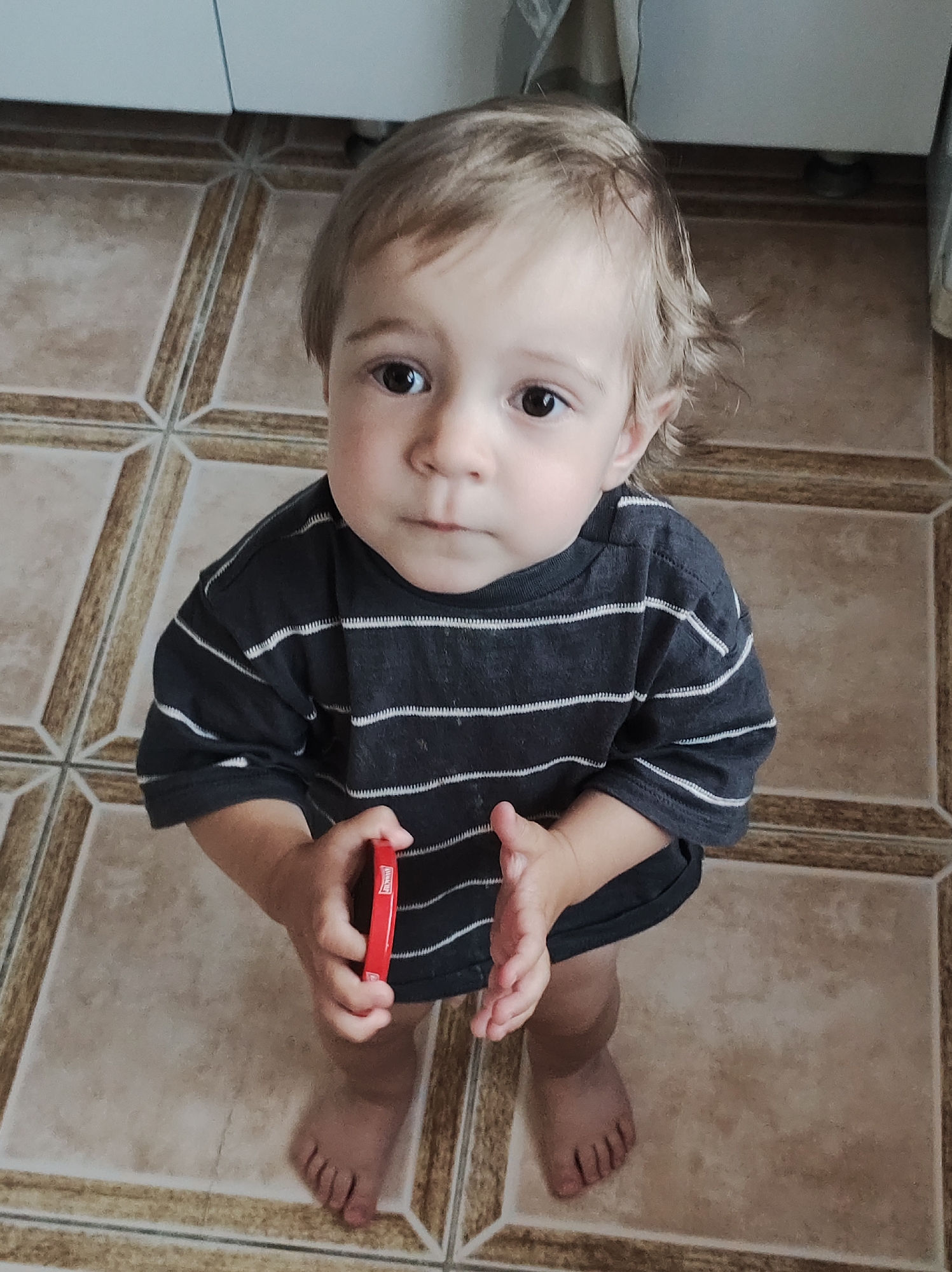 A young child, likely a toddler, is featured prominently in this high-angle indoor photograph taken in Chaikovsky, Russia. The child has light brown hair, dark eyes, and a fair complexion. They are wearing a dark grey or black short-sleeved t-shirt with thin white horizontal stripes.
The child stands barefoot on a tiled floor, which has a pattern of light beige and brown tiles, featuring a distinctive star-like design where four tiles meet. In the background, parts of light-colored, possibly white, cabinets or furniture are visible, suggesting the setting is a kitchen or a room with built-in storage.
The child is looking directly up at the camera with a serious or curious expression. Their hands are clasped in front of them, and they are holding a small, red, rectangular object. Faint white text is visible on this red object, which appears to read "MINI-B". There are no explicit cues for the time of day, but the indoor lighting suggests daytime.