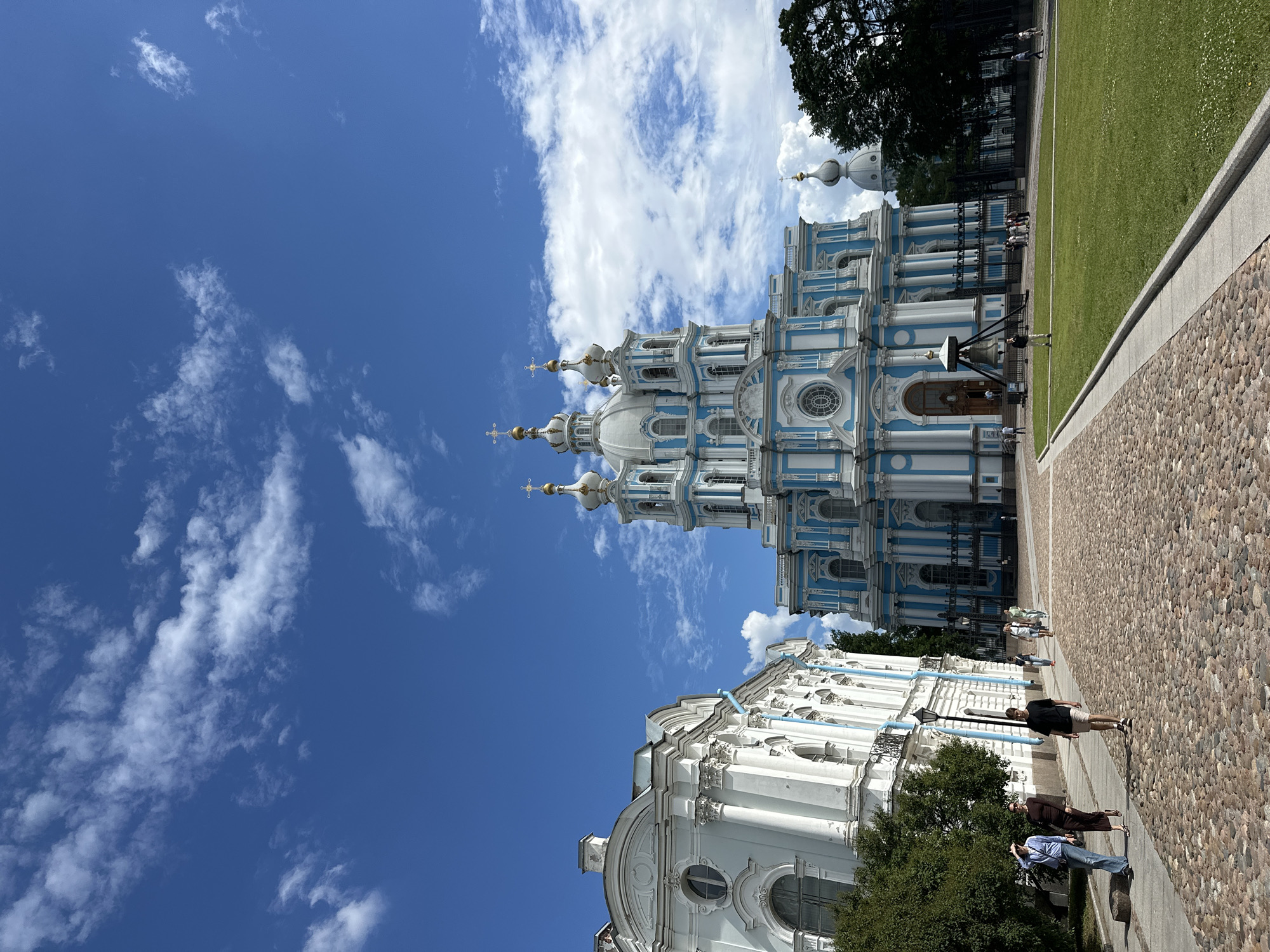 The image shows the Smolny Cathedral in Saint Petersburg, Russia, on a bright, sunny day with scattered clouds. The cathedral is a prominent blue and white Baroque building with multiple domes. To the left of the cathedral is another white building with similar architectural features. In the foreground, a cobblestone path leads towards the cathedral, and a green lawn is to the right. Several people are walking on the path, some strolling and others appearing to be tourists. The sky is a vibrant blue, with wispy white clouds adding texture and depth to the scene. Trees are visible on the right side of the image, and a fence surrounds part of the cathedral grounds.