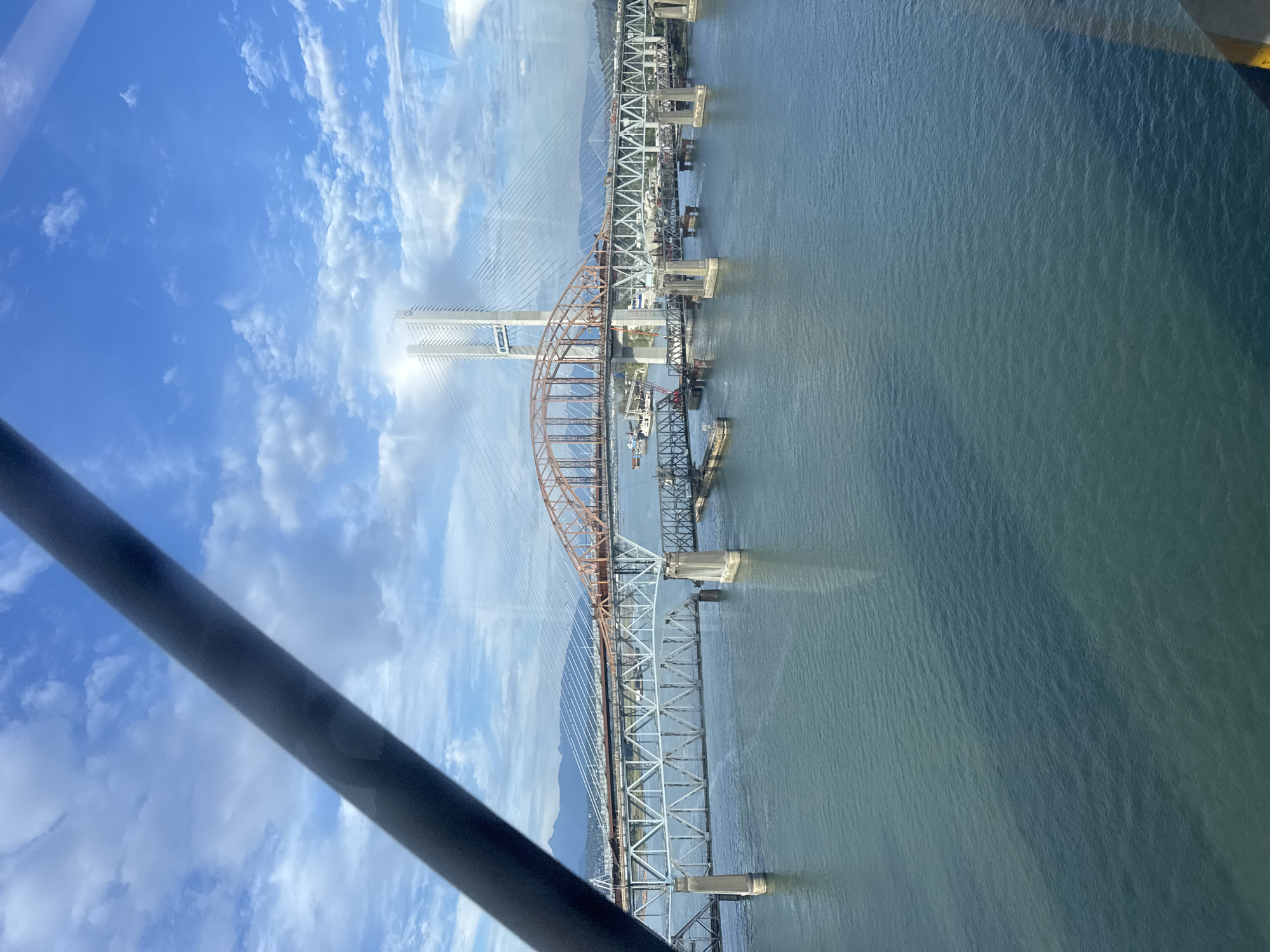 The image shows a view of the Fraser River in New Westminster, Canada, featuring multiple bridges. In the foreground, the choppy, dark blue water of the river is visible. Several bridges span the river, including a steel truss bridge with concrete piers and a rust-colored arch bridge. In the background, a modern cable-stayed bridge with tall white towers is prominent. The sky is a bright blue with scattered white clouds. The overall scene depicts an urban riverfront with significant infrastructure.