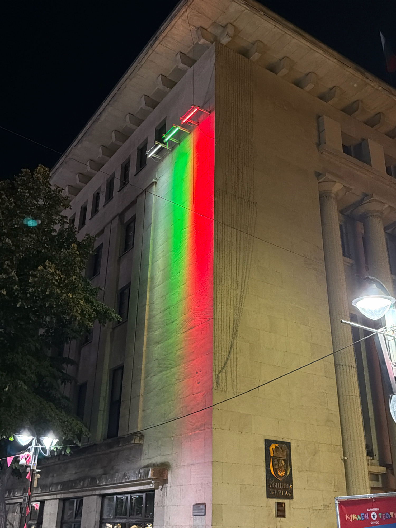 The image shows a building facade at night, illuminated by a vertical band of green and red light projected onto its side. Two fluorescent light fixtures are mounted horizontally near the top of the illuminated section, emitting the colored light. The building appears to be constructed of light-colored stone blocks and features classical architectural elements such as columns and decorative cornices. A large tree with green foliage is visible to the left of the building. Streetlights and some red and white flags are present at the bottom of the frame, suggesting a street or public area. A plaque with a coat of arms and text in Cyrillic script, reading "ОБЩИНА БУРГАС," is affixed to the building's facade, indicating the location is Burgas Municipality in Burgas, Bulgaria. Below this, a partial view of a red banner with Cyrillic text is visible, potentially advertising an event. The overall scene is a nighttime urban view of a municipal building adorned with colorful light projection.