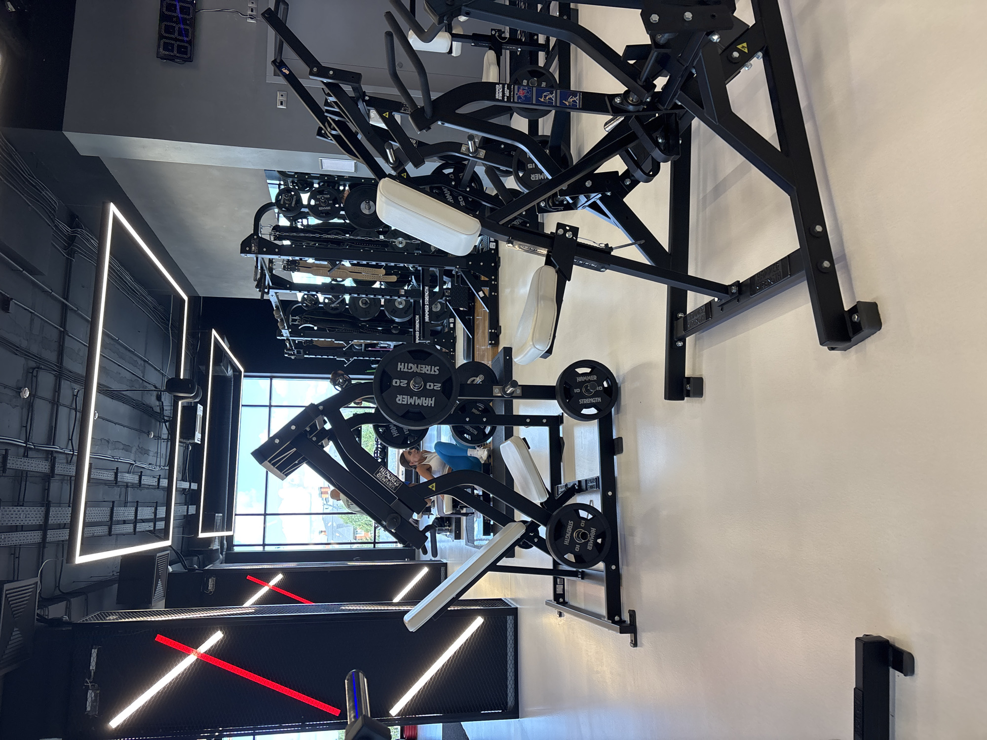 The image depicts the interior of a modern, well-lit gym, specifically located in Monolit, Russia. The foreground features a black and white weight machine, possibly a leg press, with white padded seating and footrests. On the machine are several black weight plates, some labeled "HAMMER STRENGTH 20 kg". In the mid-ground, another similar weight machine is visible, with a young woman wearing a white sports bra and blue leggings sitting on it, appearing to be engaged in a workout. She is looking towards the machine. Behind her, rows of weight racks filled with various weights and gym equipment are arranged. The gym has a high ceiling with exposed industrial elements, including ventilation systems and conduits, illuminated by long, rectangular LED light fixtures and striking diagonal red and white LED strips on a black wall. A large digital clock displaying "88:88" is mounted on a grey wall in the background. Large windows offer a view of the exterior, showing a building and trees under a bright sky. The floor is a light-colored, seamless surface.