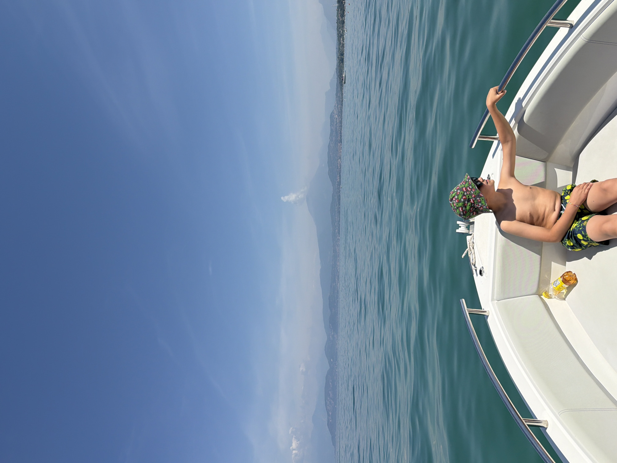 A young boy sits on the front of a white boat in the middle of a body of water. He is wearing a colorful hat and sunglasses, with his left arm resting on the boat's railing. A bottle of yellow liquid is on the seat next to him. The water is a deep turquoise, and the sky is a clear blue. In the distance, a hazy mountain range lines the horizon. The image appears to have been taken on a sunny day. The location is Castelnuovo del Garda, Italy.