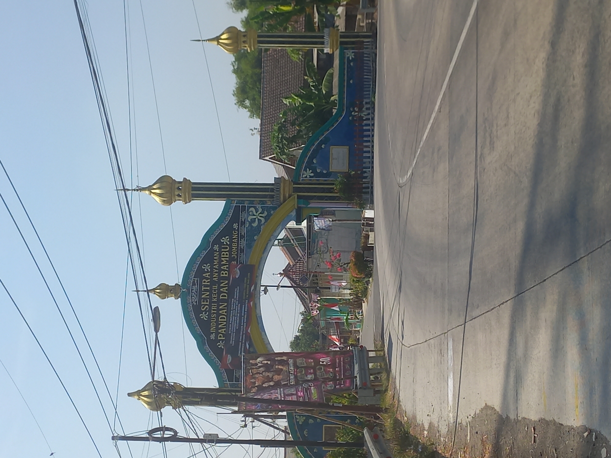 The image captures a street scene in Genengan Jasem, Indonesia, dominated by a large, ornate archway. The archway is painted in green and blue with gold embellishments and features Islamic architectural elements like minaret-like towers topped with golden domes. The text on the arch reads "SENTRA INDUSTRI KECIL ANYAMAN PANDAN DAN BAMBU B. JOMBANG," indicating that this is a center for small-scale industries related to woven pandan and bamboo, located in the Jombang regency. A large banner hangs from the arch on the left side, displaying images of people and text, possibly advertisements or information about local events or businesses.
The road in front of the archway is paved and shows tire marks and faint white lines. To the left of the road, a sidewalk area is visible with some litter and vegetation. Beyond the archway, a few buildings with tiled roofs can be seen, along with trees and more vegetation. The sky is clear and blue, suggesting a sunny day. Electrical wires are strung across the upper part of the image, supported by poles. The overall impression is a local gateway marking an area dedicated to handicraft production.