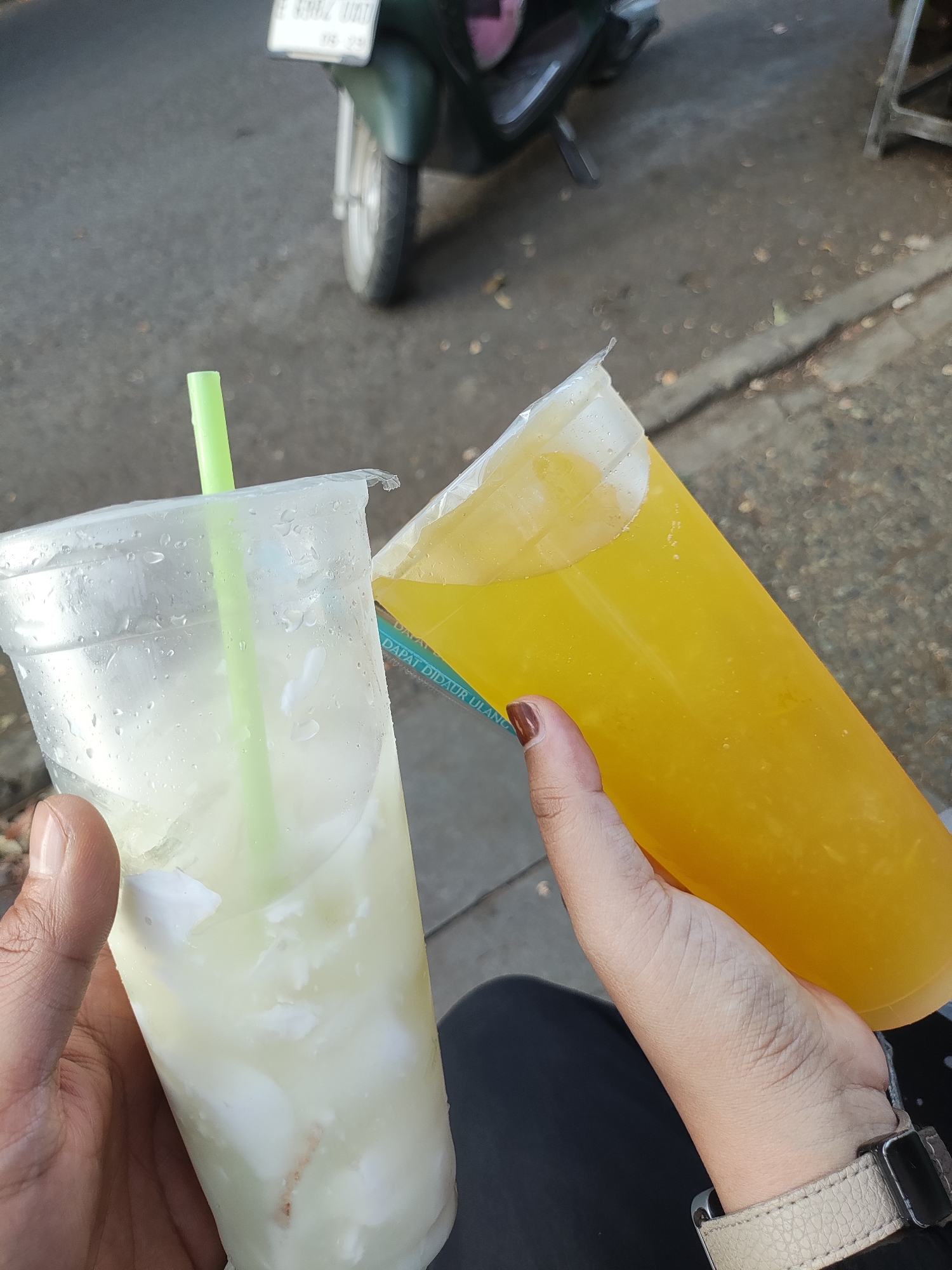 The image shows two hands holding drinks. The left hand, a male's, holds a clear plastic cup filled with a white, cloudy liquid containing chunks of white material, possibly coconut flesh. A green plastic straw protrudes from the cup. The right hand, a female's, holds a larger, clear plastic cup filled with a bright yellow liquid. The cup has a white lid. A faint blue text, "DAPAT DIDAUR ULANG," which translates to "RECYCLABLE," is visible on the side of the yellow drink's cup. The background suggests an outdoor urban environment, with a paved road, some fallen leaves, and a portion of a green motorcycle visible. The overall scene is consistent with a street vendor or casual outdoor setting in Cirebon, Indonesia.
