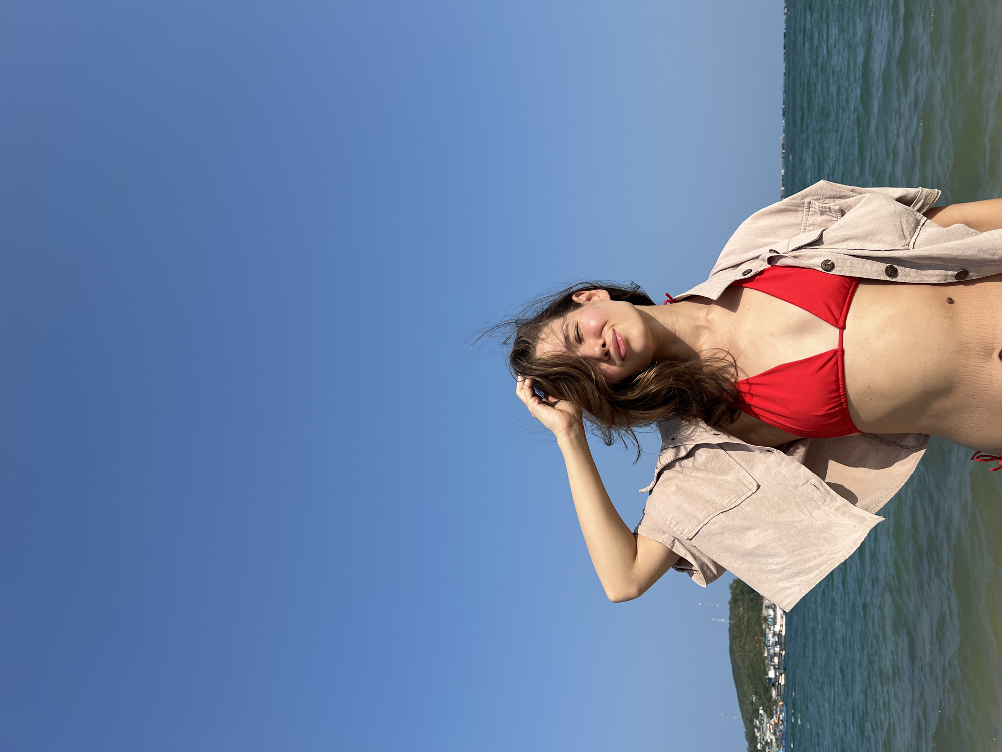 A young woman is captured posing outdoors against a backdrop of a clear blue sky and the ocean, indicative of a coastal setting like Nam Nha Trang, Vietnam. She is positioned on the right side of the frame (when correctly oriented), looking towards the viewer with her head tilted slightly upwards and to the left. Her long, dark brown hair frames her face, and her right hand is casually raised, touching her hair. She is wearing a bright red bikini top, covered partially by a light-colored, unbuttoned shirt or jacket. Her expression is relaxed, with her eyes partially closed and a subtle smile, suggesting a serene or playful mood.
The upper and left portions of the image are dominated by a vast, cloudless blue sky, indicating bright, sunny weather. To the right, the ocean stretches out, showing gentle waves and meeting a distant landmass on the horizon. This landmass features various buildings, characteristic of a developed coastal area. The bright illumination confirms it is daytime, likely mid-day or early afternoon. The overall scene conveys a feeling of summer and relaxation by the sea. There is no visible text in the image.