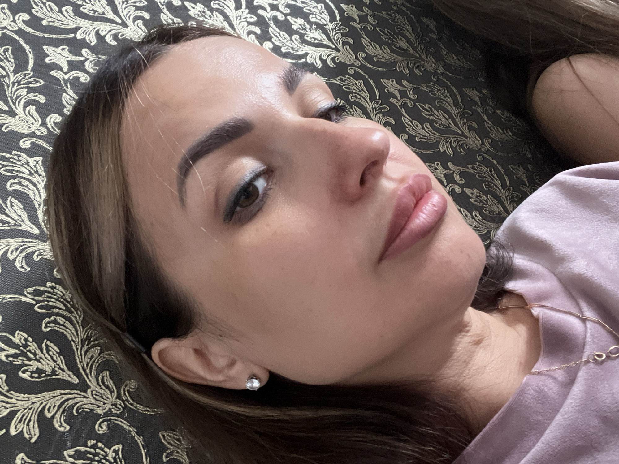 A close-up, eye-level medium shot captures the face of a woman in her late 20s or early 30s, angled to her right. She has dark brown hair with lighter streaks, pulled back and partially obscuring her right ear, which is adorned with a diamond stud earring. Her makeup includes defined eyebrows, winged eyeliner, and subtle eyeshadow. The woman is wearing a light purple long-sleeved shirt with a delicate gold necklace featuring two small loops. Her lips are slightly parted, showing a hint of her teeth. The background is a dark, textured wallpaper with a repeating pattern of gold floral motifs. The lighting is soft, highlighting the contours of her face and casting subtle shadows.