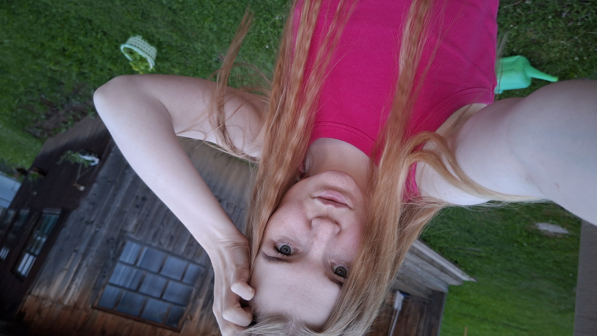 A selfie-style portrait captures a young person, likely female, lying on their back outdoors, with the image rotated 90 degrees clockwise. The individual has fair skin, light-colored eyes, and long, flowing reddish-blonde hair that frames their face. They are wearing a bright pink ribbed tank top and look directly into the camera with a neutral, calm expression. Their left arm is bent, with the hand resting near their head.
The background suggests a casual outdoor setting in Bolshoy Bukor, Russia. Lush green grass covers the ground, indicating a yard or garden. To the upper left, a light-colored woven basket is partially visible on the grass. In the upper right, a bright green plastic watering can sits on the ground. A rustic, dark wooden building, possibly a shed or an older house, occupies the left and lower portions of the scene, featuring multiple windows that show some reflections. The lighting appears soft and natural, suggesting it could be late afternoon or early evening. There is no visible text in the image.