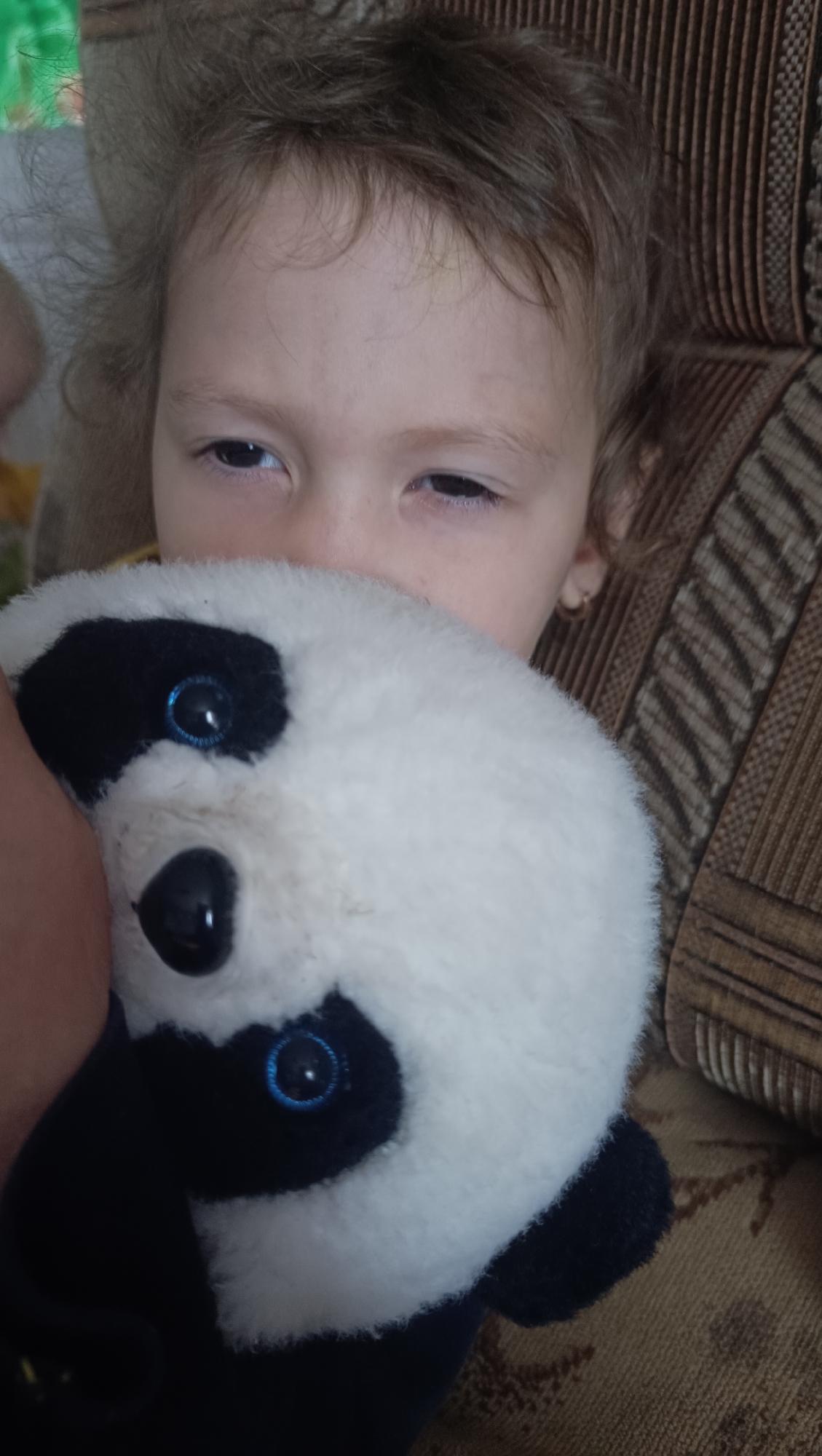 In this close-up, head-and-shoulders shot, a young child, appearing to be around 4-6 years old, is nestled against a plush panda toy. The child's face is partially obscured by the panda, but their eyes are visible, looking slightly downwards with a gentle, sleepy expression. The child has light brown, slightly tousled hair and is wearing small gold hoop earrings. The panda toy is white and black, with large blue eyes and a black nose. The background consists of a textured, patterned sofa in shades of brown and beige, suggesting an indoor, domestic setting. The lighting appears to be natural, coming from the side, casting soft shadows. The image conveys a sense of comfort and quiet intimacy, with the child holding the panda close. There is no indication of the city of Karsha, Russia, or any elements that would place the scene geographically.