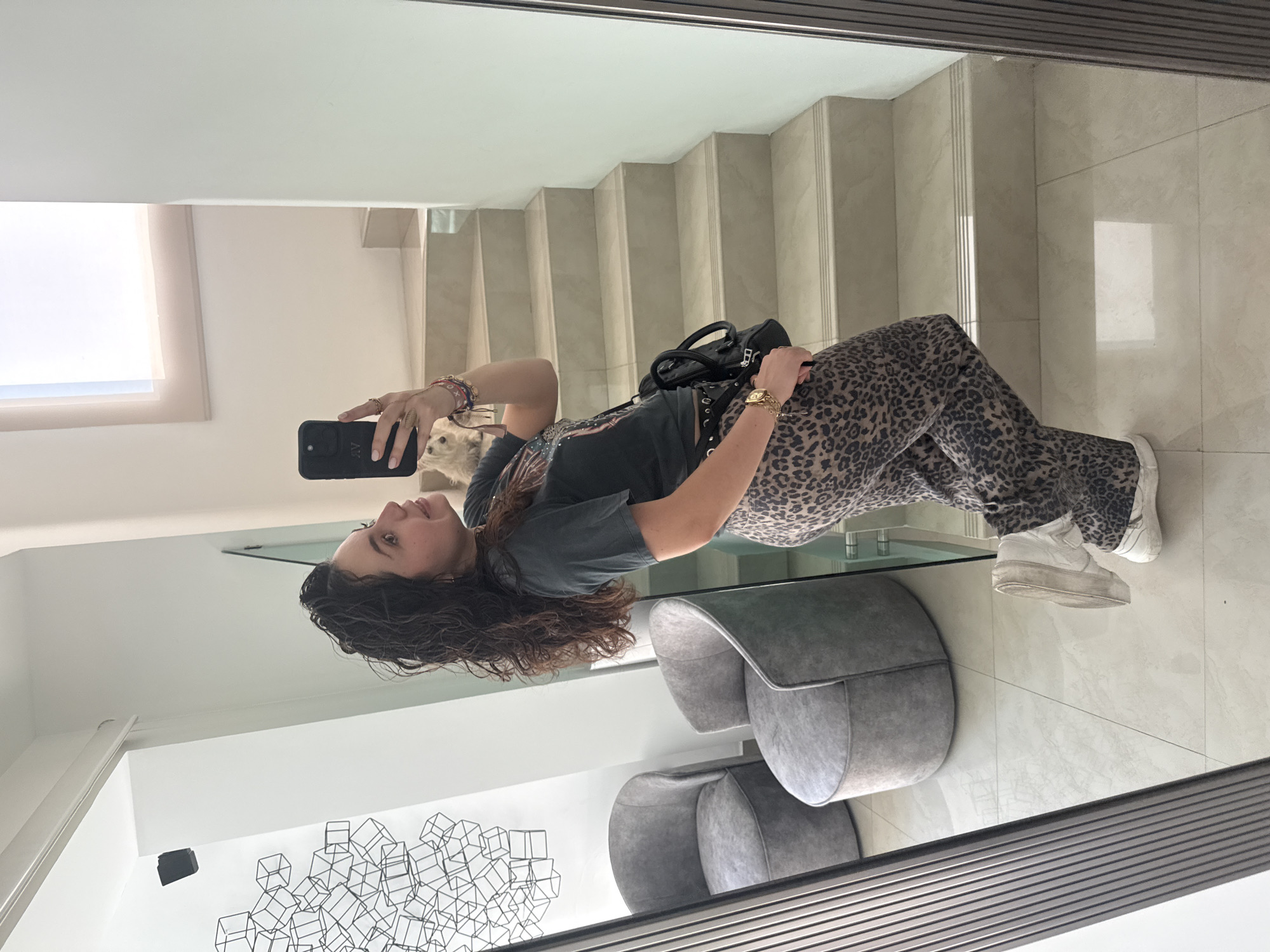 A woman stands in front of a full-length mirror taking a selfie with a black smartphone. She has long, curly brown hair and is wearing a dark graphic t-shirt, leopard-print pants, and white platform sneakers. A black handbag is slung over her shoulder. The room is modern with a staircase in the background. Two gray, rounded chairs are visible, along with a decorative art piece made of black wire cubes on the wall. The floor is tiled in light gray. A small, white dog is partially visible near the woman's arm. The location appears to be indoors. The location within Santa Clara Ocoyucan, Mexico cannot be determined based on the image.