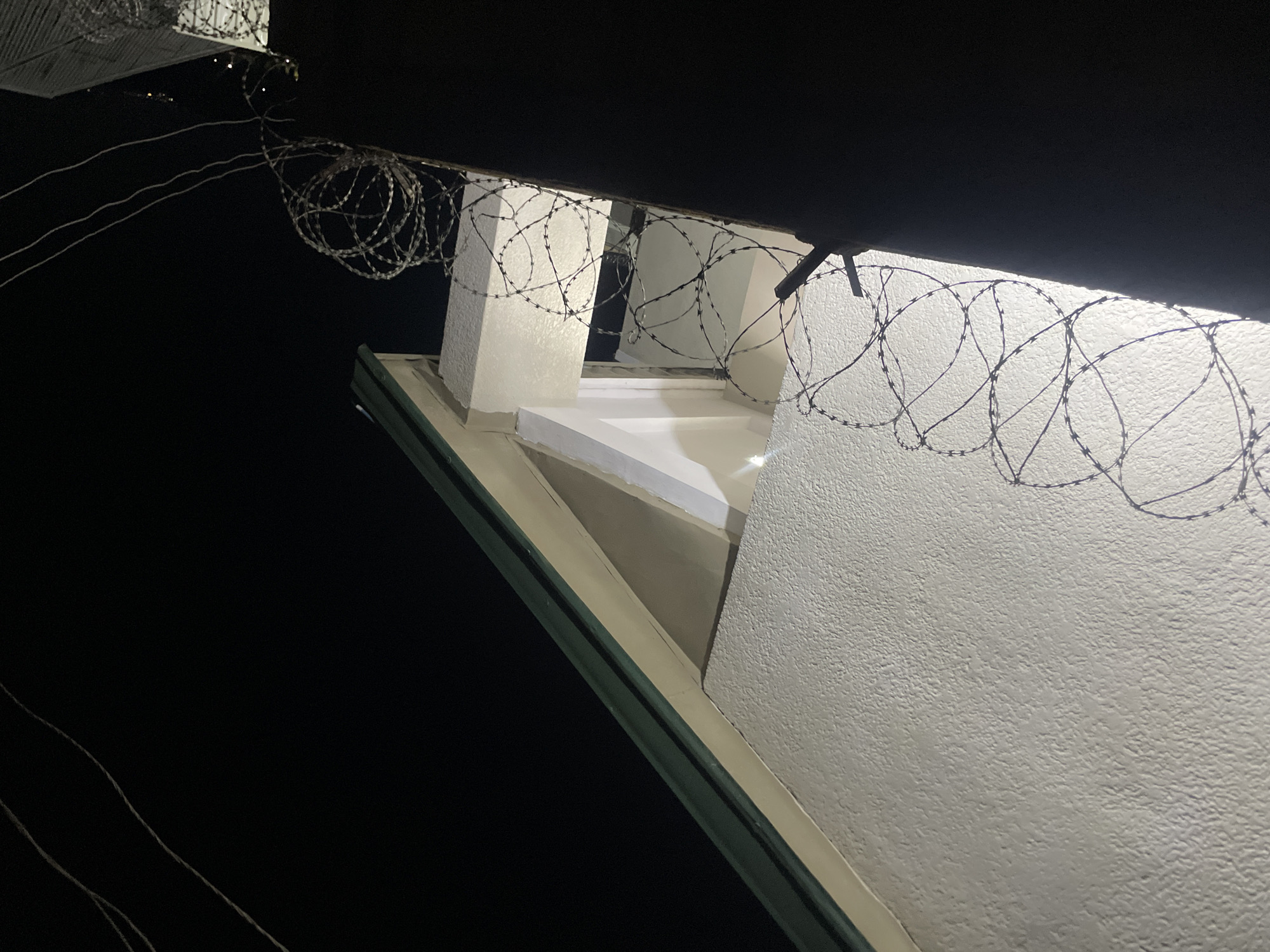 This image depicts a night scene in Jos, Nigeria, focusing on a building's exterior, likely a security perimeter. The foreground is dominated by a dark, textured wall or fence topped with coiled barbed wire, suggesting a secure or restricted area. Behind the barbed wire, a portion of a building is visible, characterized by a rough, textured white wall and a protruding gable with a green roof edge. An opening or window is visible within the gable structure, dimly lit from within, casting a soft light on the surrounding architectural elements. The background is a dark, starless night sky. Several thin wires, likely utility lines, are visible against the dark sky in the upper right portion of the image. The overall impression is one of security and nighttime visibility.