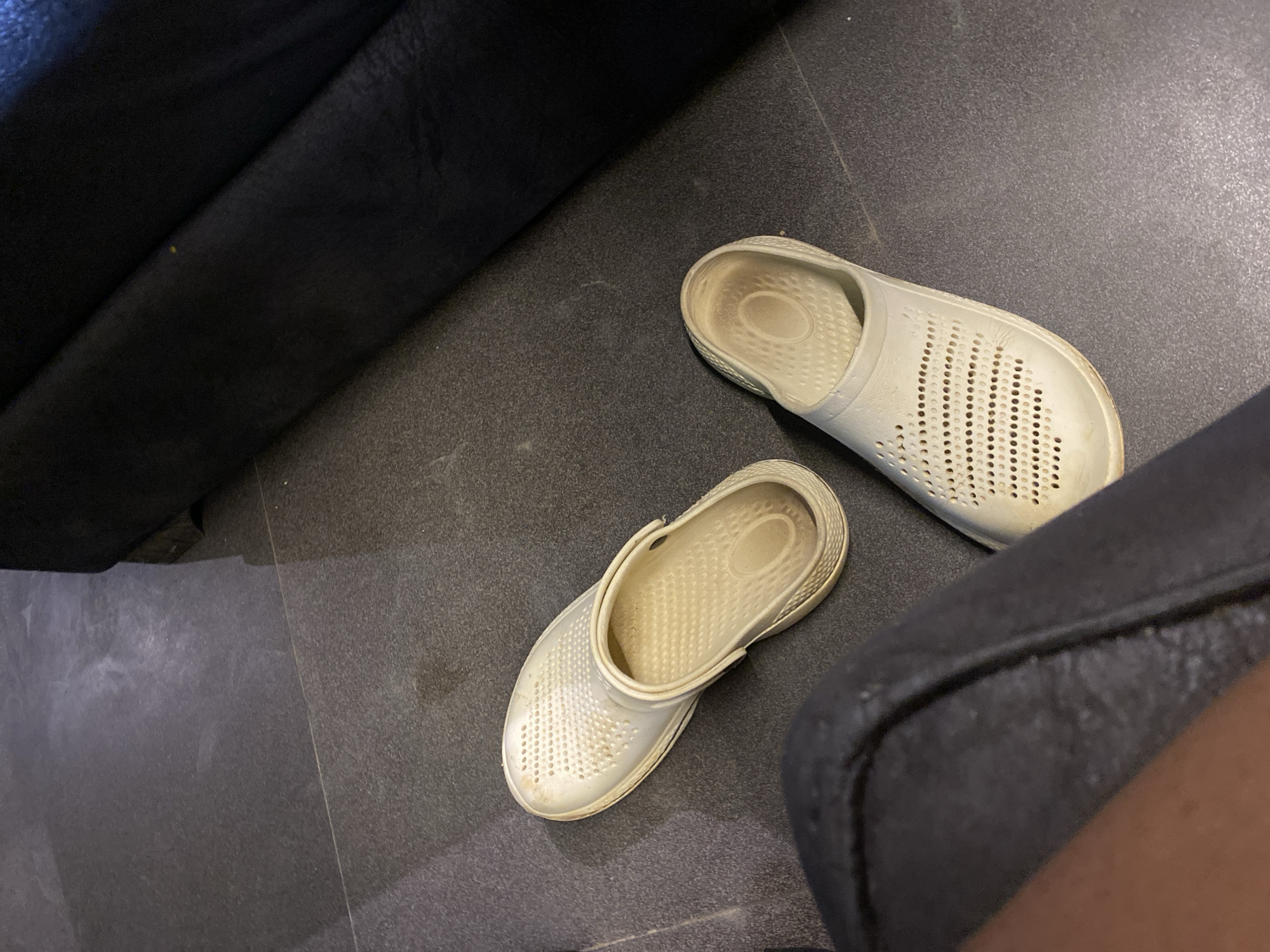 A pair of white clogs are on a dark gray tiled floor. One clog is on its side, showing the interior. The other clog is upright. To the right, a dark, upholstered surface, possibly furniture, is partially visible. A dark fabric, possibly clothing, is in the lower left corner of the frame. The scene appears to be indoors, in a residential or commercial setting. The tiled floor has faint lines indicating grout. There is a slight sheen on the tiles, suggesting they are polished. There are no people visible in the image, and no activities are depicted. The lighting is dim, casting soft shadows. The image is taken from a high angle.