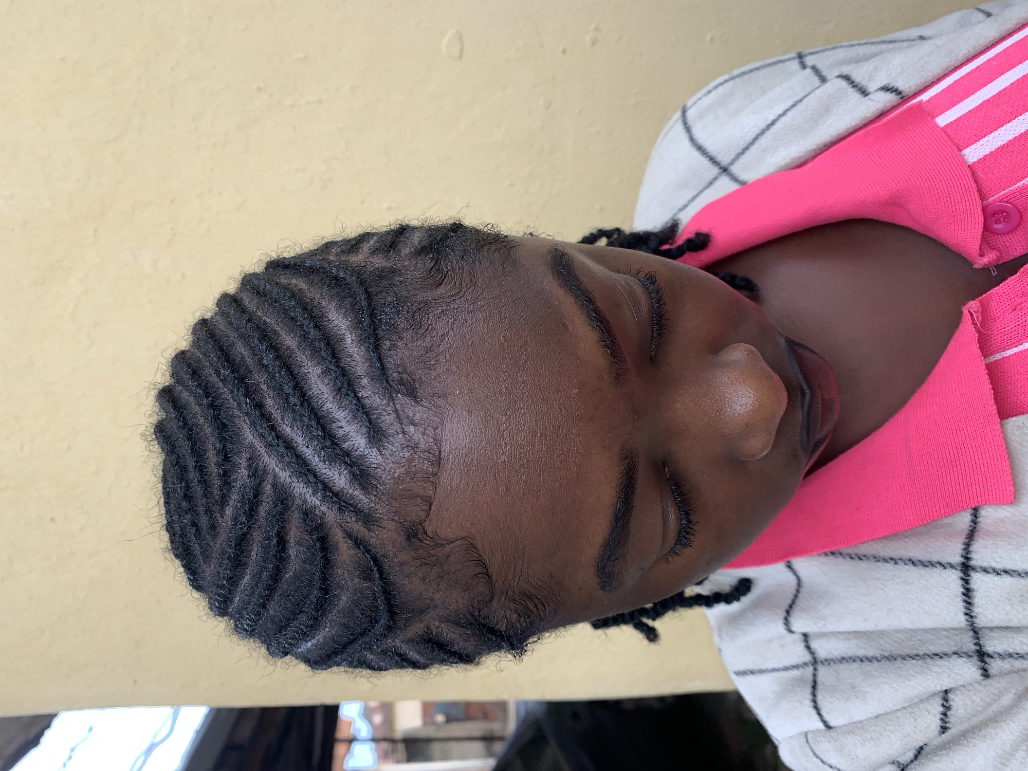 In the city of Rafin Par, Nigeria, a close-up, slightly overhead shot captures an individual with dark skin, looking downwards. The person's hair is styled in approximately eight neat cornrows, braided in a distinct wavy or zig-zag pattern across the scalp from front to back. Edges are laid and styled around the forehead. The individual's eyes are mostly closed, and a portion of their forehead, nose, and lips are visible. They are wearing a vibrant fuchsia pink collared shirt, featuring subtle vertical white stripes on the placket, under a white garment with a dark, possibly black, checkered pattern. The background consists of a textured, plain light yellow wall. In the upper left corner, out-of-focus elements suggest an outdoor setting, including a blurry structure and faint outlines of what might be a vehicle.