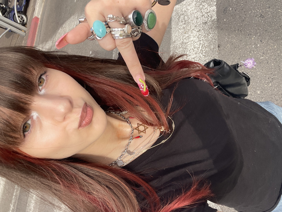 A young adult female is depicted in a close-up, upward-angle front-facing image, captured outdoors in Raanana, Israel. She has brown hair with red streaks and bangs, hazel eyes highlighted with white eyeliner, and her lips are slightly pursed. She wears a black t-shirt, light blue jeans, and carries a black shoulder bag. Her neck is adorned with multiple layered necklaces, including a prominent silver-colored chain with a Star of David pendant and red chili charms. Her right hand is raised with the index finger extended, displaying numerous silver-colored rings: a large light blue stone on the thumb, various intricate bands on the index finger, a dark green stone on the middle finger, and a light green stone on the ring finger. Her fingernails are long and sculpted, with the thumb nail painted pink and the index finger nail red with yellow-green accents. The background features a paved surface with white markings, consistent with an urban road or crosswalk, and a blurred dark vehicle in the distance.