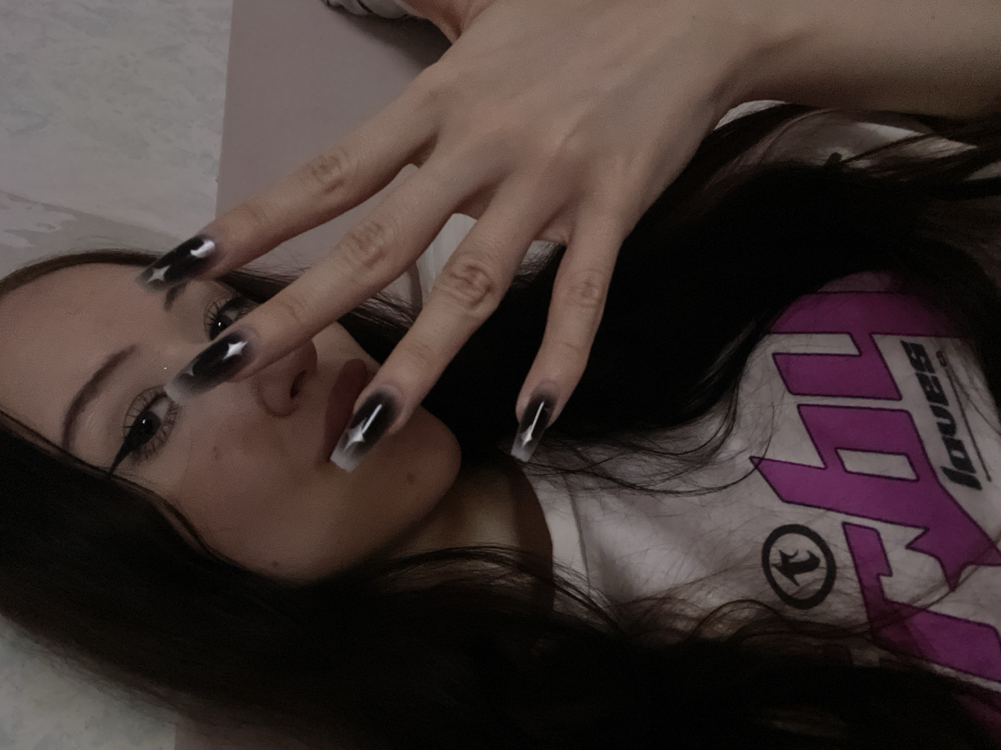 The media file is a close-up, low-angle selfie of a person lying down, with their face partially obscured by their hand and dark hair. The person has long, dark hair and is wearing a white t-shirt with a large, stylized purple logo. The logo features sharp angles and the partial word "LOVES" is visible. The person has dramatic eye makeup with winged eyeliner and is wearing long, pointed artificial nails with a black and white gradient design, featuring small white star or arrow details. The lighting appears to be artificial and dim, suggesting an indoor setting at night. The scene conveys a relaxed or intimate mood. There are no direct indicators of the time of day or weather, but the indoor setting and lighting suggest it's not daytime outdoors. No specific location cues are visible to pinpoint a location within Votkinsk, Russia, beyond the general context provided.