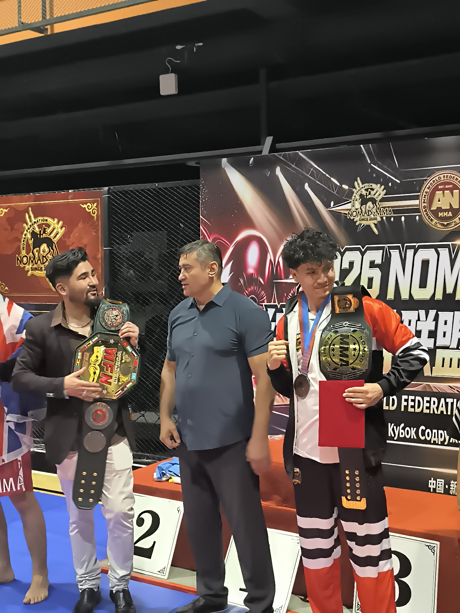 Three men are posing for a photograph at what appears to be a sporting event. Two men are wearing championship belts. The man on the left is wearing a suit jacket and holding a belt. The man in the middle is wearing a blue shirt and looking at the man on the left. The man on the right is wearing a track jacket and holding a belt and a red folder. There is a backdrop with event logos and text in English and other languages. The scene is brightly lit, likely indoors under artificial lighting.