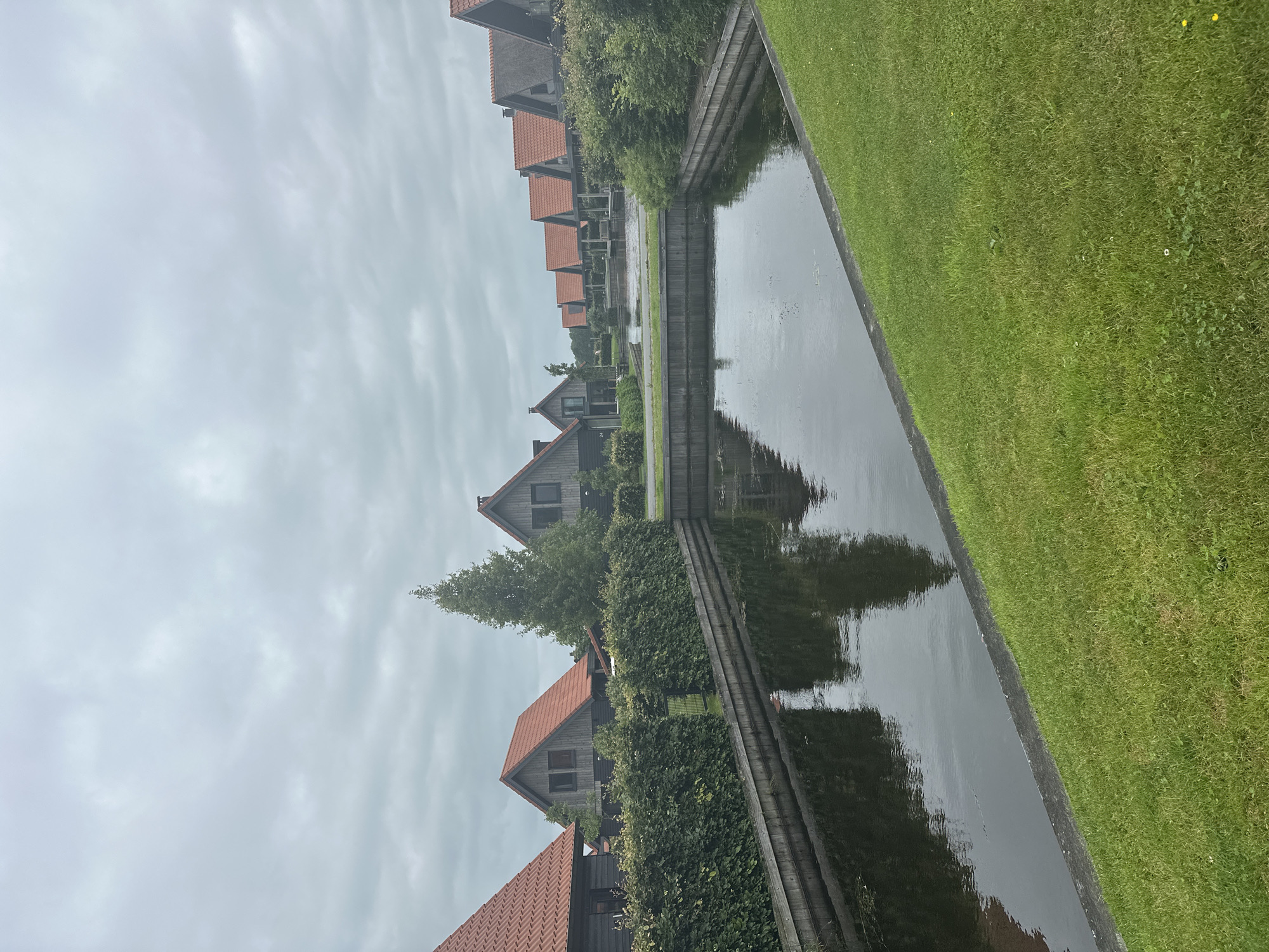 This is a medium shot, eye-level, exterior, vertical photograph of a row of modern holiday homes with red tile roofs in Ossenzijl, Netherlands. The homes are dark wood with grey trim, and they line a canal that runs along the bottom of the image. The canal is bordered by a wooden retaining wall and a hedge. Green grass slopes down from the right side of the frame towards the canal. The sky is overcast and grey, with scattered clouds. There are no people visible in the image.