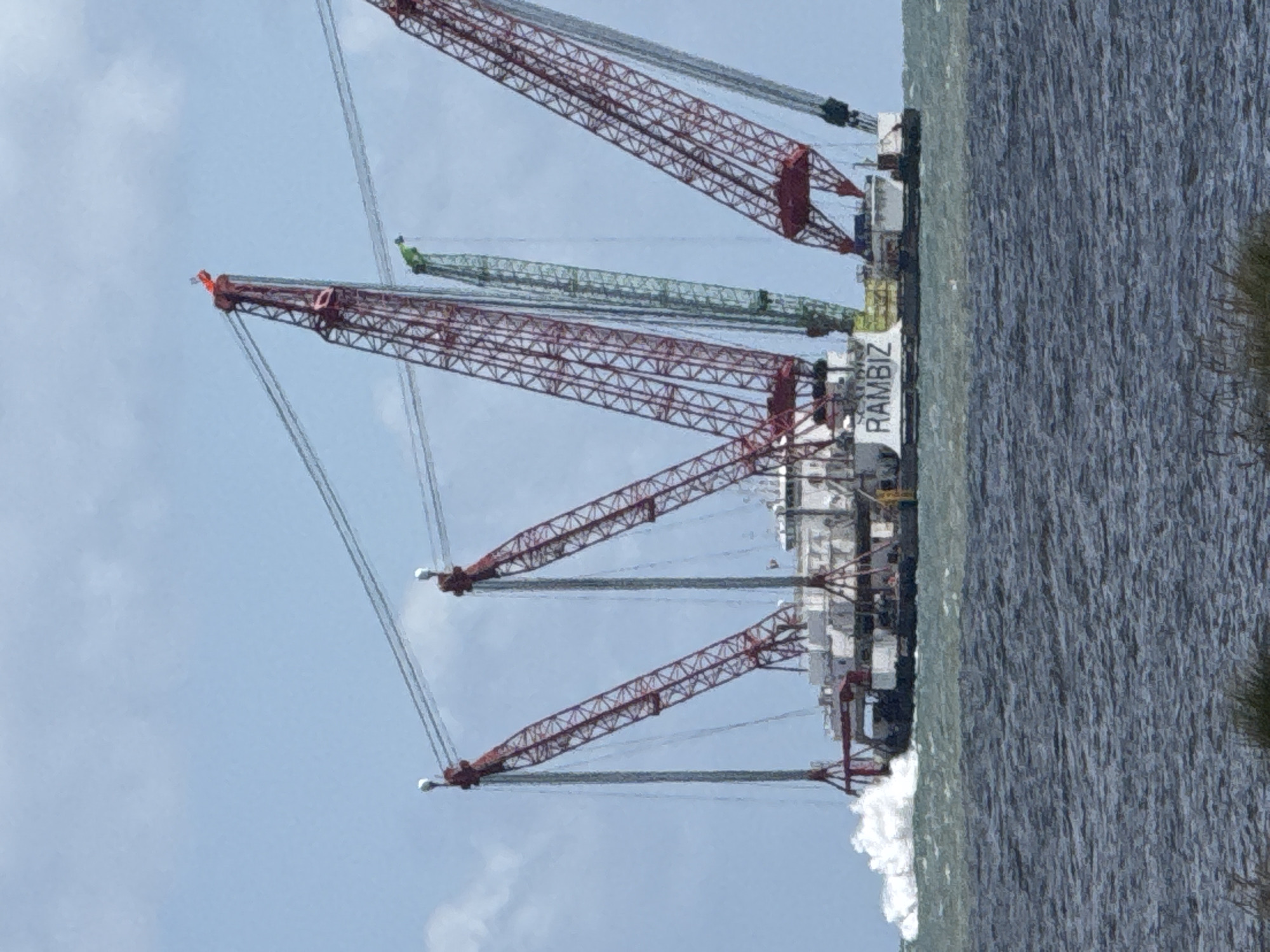 The image shows a large, offshore vessel named "RAMBIZ" equipped with multiple cranes, including one green crane, positioned in water near what appears to be a shoreline. The vessel is near the city of Great Yarmouth, United Kingdom. The cranes are various shades of red and grey. The sky is light blue with sparse clouds. The sea has visible wave action. Some green vegetation can be seen on the right edge of the image.