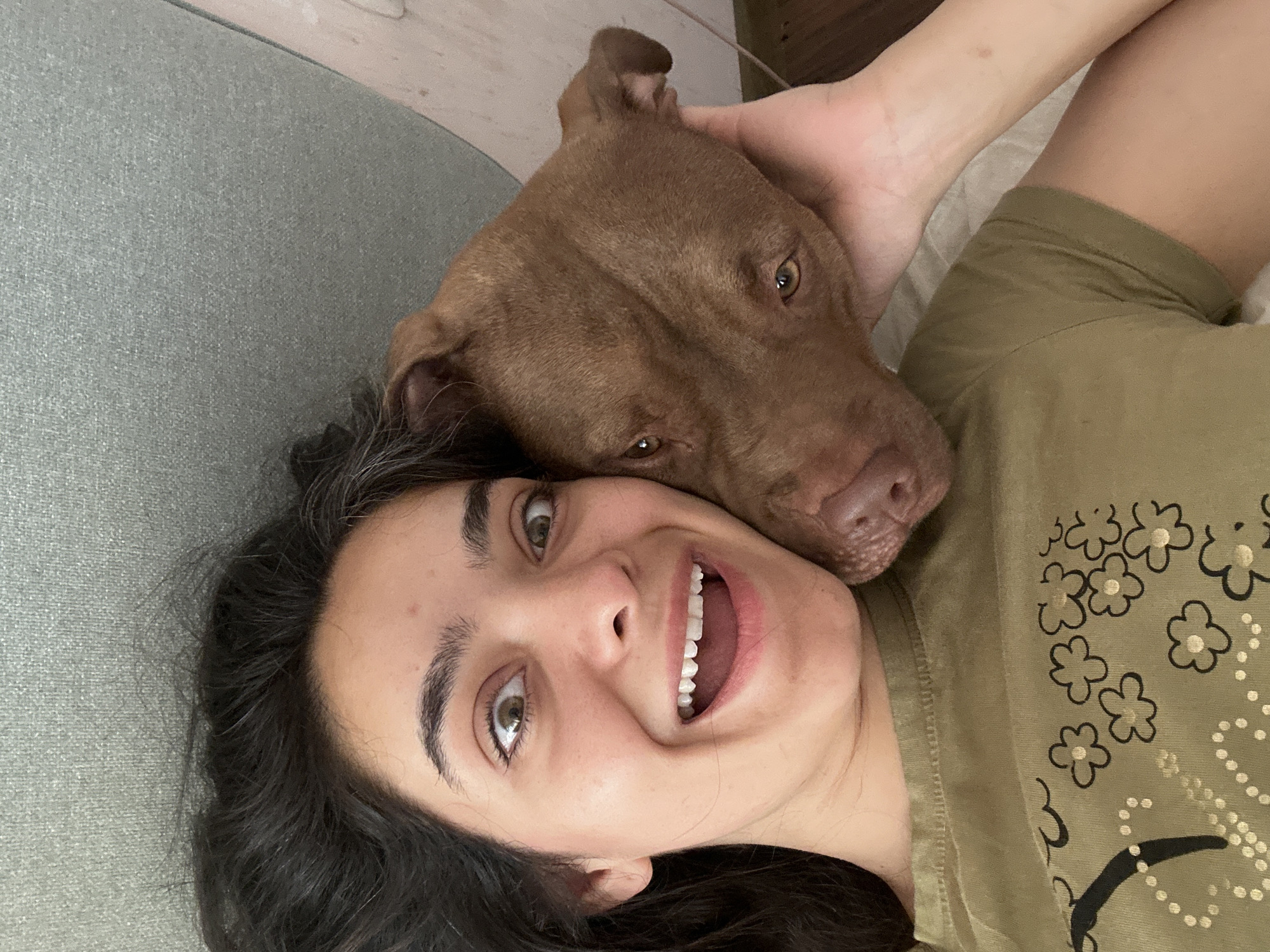 A woman and a brown pit bull dog are lying down together. The woman is on the left, looking directly at the camera with a surprised expression, her mouth open in a wide smile, and her eyes wide. Her dark hair is spread out around her head. The dog's head is resting on her right cheek and neck, with its brown eyes looking forward. The dog's left ear is slightly floppy. The woman is wearing a green t-shirt with a floral pattern. The background shows a grey upholstered surface, possibly a sofa, and a hint of a pink wall on the right. The scene is lit with natural light. This image was taken in Malevo, Bulgaria.