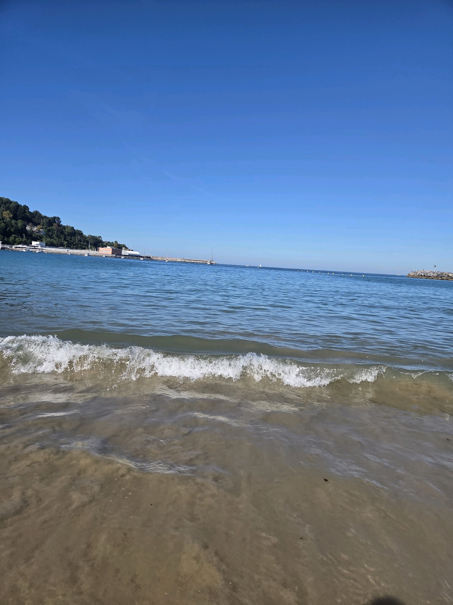 This is a horizontal, eye-level, medium shot of a sandy beach and the sea under a clear blue sky in Hondarribia, Spain. In the foreground, a small wave crashes on the wet sand, with the shallow water appearing murky. The sea extends to the horizon, with several buoys visible in the distance. On the left side of the image, a tree-covered hill rises, with buildings and a pier situated at its base. A breakwater made of large rocks extends into the sea on the right side of the frame. The overall scene depicts a calm, sunny day at the coast.