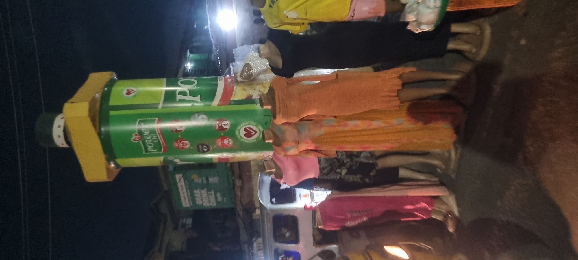 The image depicts a nighttime street scene in Lagos, Nigeria. Multiple mannequins are displayed in a row, possibly for a clothing vendor, on what appears to be a sidewalk or beside a road. They wear various styles of clothing: a yellow top, a black dress, an orange dress, a tie-dye dress, a pink top with a black skirt, and a pink top with words on it. Behind the mannequins is a white van. A prominent advertising display featuring "Power Oil" stands out. The overall lighting suggests the scene is illuminated by streetlights and the headlights of vehicles.
