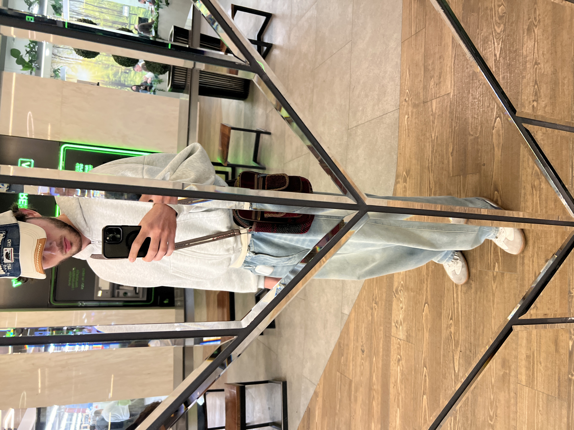 The image shows a person taking a selfie in front of a mirrored display unit. The reflection shows a male wearing a grey hoodie, light blue jeans, and white sneakers, with a crossbody bag. He is holding a black smartphone. Reflected behind him is an ATM and a wall with plants visible. The background suggests an indoor setting, likely a shopping mall or commercial space, in Yerevan, Armenia.