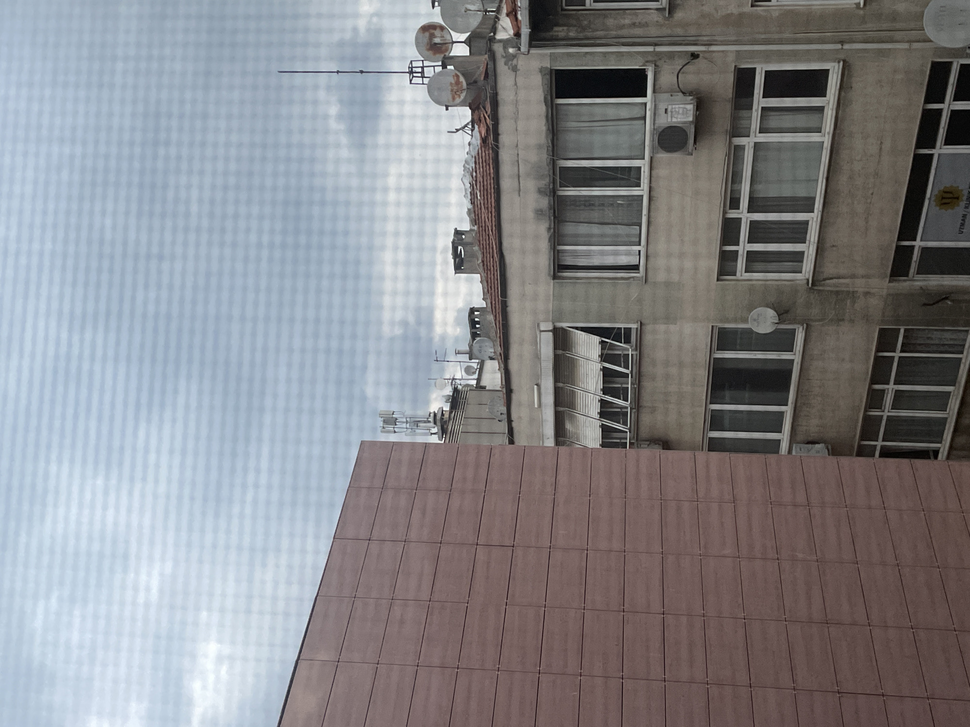 The image depicts an exterior view from a window overlooking urban buildings. In the foreground to the left, a large, modern building with a reddish-brown tiled facade dominates the left side of the frame. To the right, an older, multi-story building displays weathered concrete walls, multiple windows with white frames, and various antennas and satellite dishes on its rooftop. The sky above is overcast with grey clouds, suggesting a cloudy or possibly rainy day.
The older building features several floors with rows of windows. Some windows are open, revealing interior curtains, while others are closed. An air conditioning unit is visible on one of the floors. The rooftop is cluttered with various antennas, including cell towers, and several satellite dishes, some of which appear rusty. Chimneys are also present on the roof. A sign with a yellow symbol and text, possibly a business or institution name, is visible on a window on a lower floor.
The scene is set in the city of Kadıköy, Turkey, as indicated by the presence of architectural styles common in Turkish urban areas and the specific mention of the location. The overall impression is one of a dense urban environment with a mix of older and newer construction. There are no people or animals visible in the image.