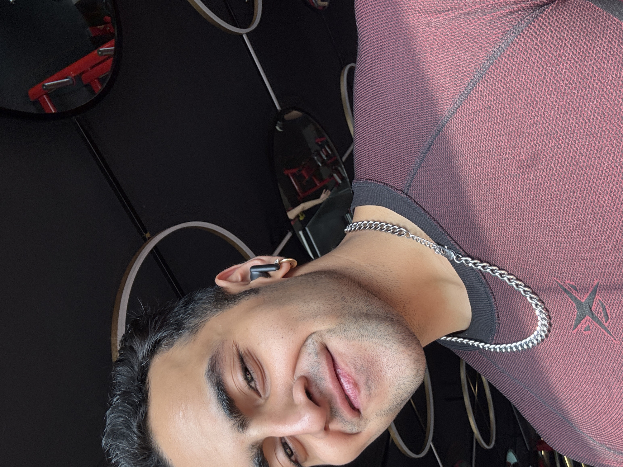 A man wearing a maroon mesh long-sleeved shirt with a black collar and silver necklace poses for a photo in what appears to be a gym or fitness center. He has short dark hair and is wearing an earbud in his left ear. A red weightlifting machine is visible in the background, reflected in a round mirror. The wall behind him is dark, adorned with decorative white circles. Given the provided location hint, the scene is hypothetically located in Kolkata, India.