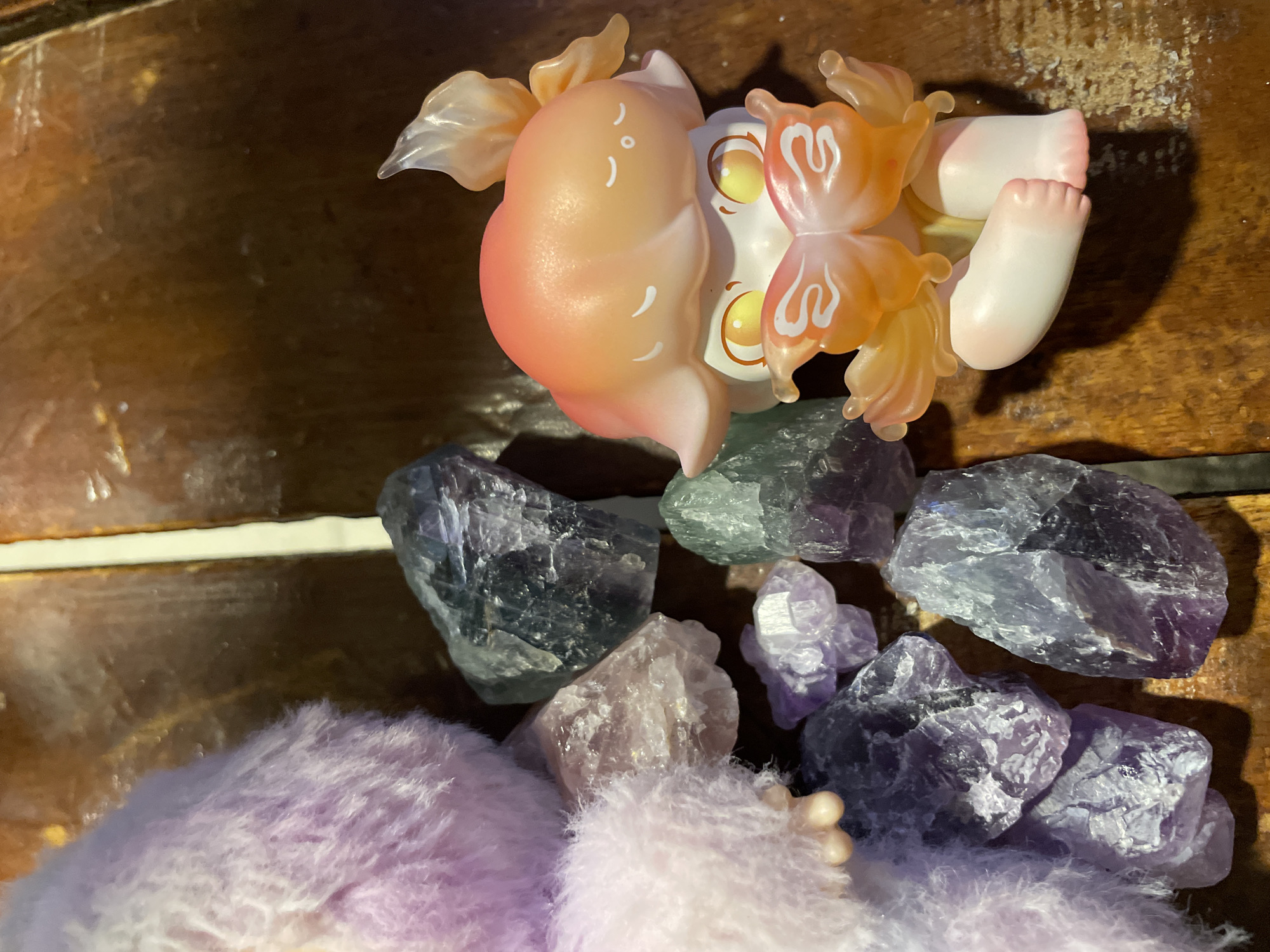 The image displays a still life composition featuring various objects arranged on a textured wooden surface. The location in the city of Davao City, Philippines, cannot be determined from the visual information provided.
On the left side of the frame, a fluffy, light purple-to-pink object, possibly a stuffed animal or a textile item, occupies a significant portion. Its texture appears soft and slightly disheveled. Adjacent to and around this object, a collection of raw, unpolished mineral specimens is scattered. These crystals vary in size and color, predominantly showing shades of translucent purple (likely amethyst), darker blue-grey, and some lighter, nearly clear or pale pinkish pieces. Their surfaces exhibit natural facets and rough textures.
On the right side of the image, a small, stylized figurine is depicted sitting. This character features a smooth headpiece with an orange-to-pale-pink gradient, adorned with small white markings suggesting closed eyes and a tiny mouth. Its face is pale, with large, prominent, glowing yellow-orange eyes. Attached to the figurine's chest are large, translucent orange butterfly wings with white decorative patterns, and additional wing-like structures are visible on the back of its head. The figurine's body is smooth and pale, with visible small feet.
All objects are resting on a surface composed of dark brown wooden planks or panels, which show grain patterns, some wear, and areas of lighter discoloration. The lighting suggests an indoor setting, casting subtle shadows from the objects onto the wooden surface.