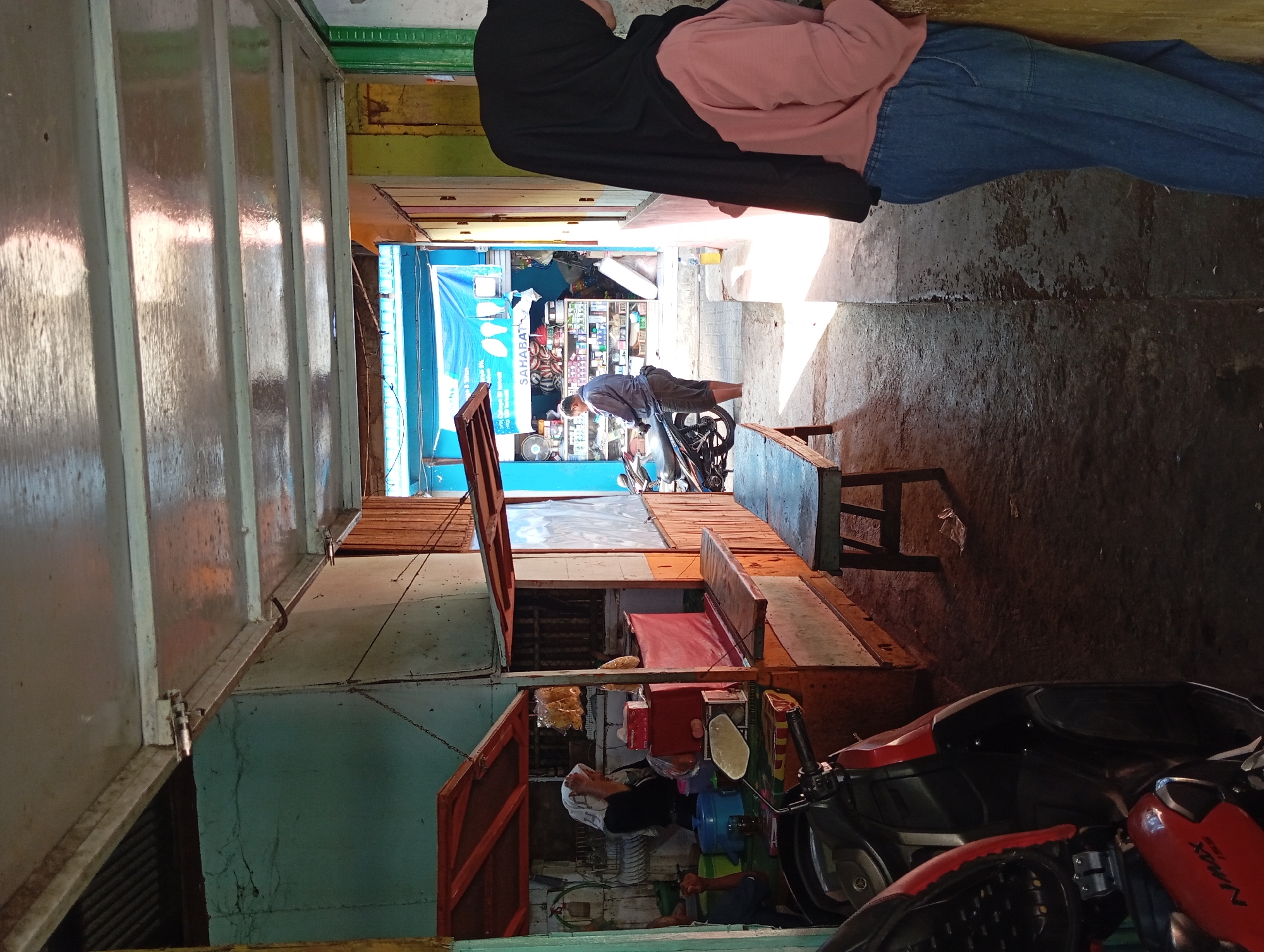 The image captures a scene in what appears to be a narrow street or alleyway in Kepulungan, Indonesia. In the foreground to the left, a red and black motorcycle is partially visible, indicating a mode of transportation common in the area. The architecture suggests a row of small shops or food stalls.
On the left side of the image, a woman wearing a white headscarf is tending to a food stall. She is positioned behind a counter where food items and containers are visible, including what look like snacks in a plastic bag and a red box. A blue plastic container and a glass are also on the counter. A second person, partially visible to the far left, is also seated.
Further down the alley, a man in a grey shirt and shorts stands next to a parked motorcycle. He appears to be looking towards a shop in the background, which is stocked with various goods, including what seem to be toiletries and food items. A blue tarp or banner with text and images is displayed above the shop.
On the right side of the alley, a person wearing a black hijab and a pink shirt is standing, leaning against a wall. They are dressed in blue jeans and are facing away from the camera, looking into the alley.
The ground is paved with concrete, and there are shadows suggesting the presence of overhead structures. A simple wooden bench is placed in the middle of the alleyway. The overall impression is of a bustling local market or commercial area with a relaxed, everyday atmosphere.