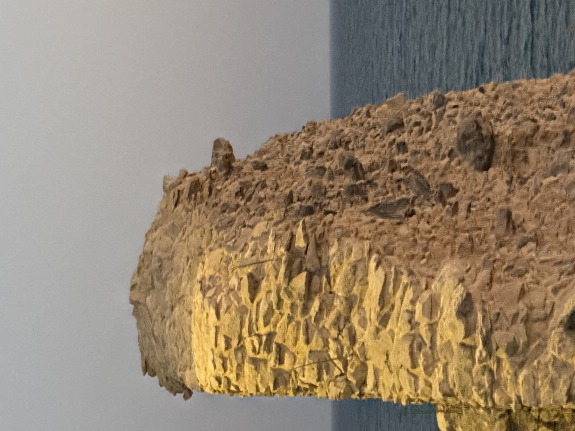The image shows a weathered stone tower standing against a pale sky and a calm blue sea. The tower is constructed from rough-hewn stones of varying sizes, appearing old and eroded. Sunlight illuminates the left side of the tower, casting warm yellow and orange tones, while the right side is in shadow. The texture of the stone is rough and uneven. In the background, the sea stretches to the horizon, with a slight haze obscuring distant details. The tower's precarious, partially ruined state suggests it is an ancient structure. This appears to be a ruin, likely a historical landmark. Given the context of Agios Tychona Community, Cyprus, this could be a section of an old fortress or watchtower, remnants of which are found in coastal areas of the island.