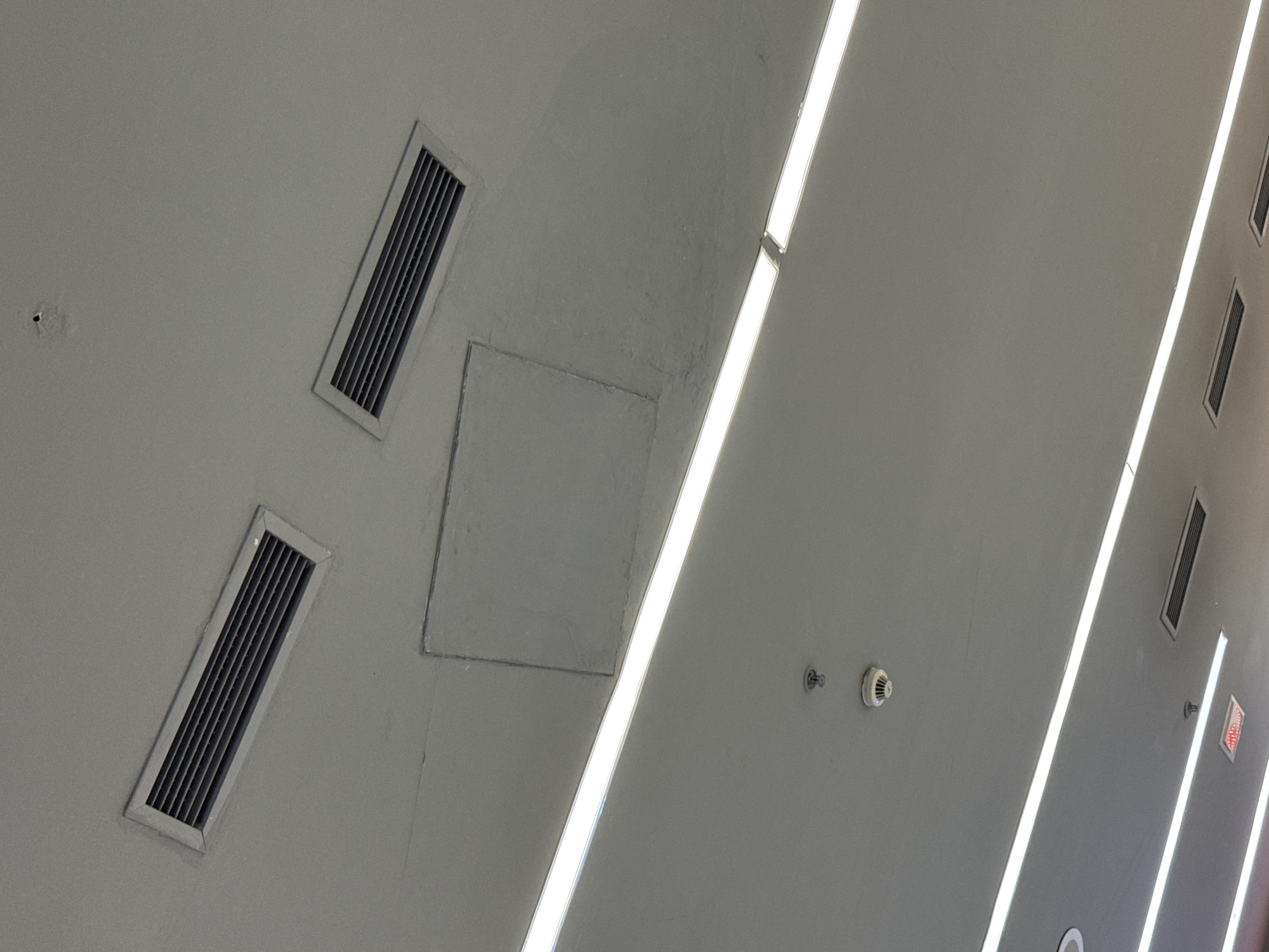 This image provides an upward view of an indoor ceiling, primarily rendered in a cool grey or off-white tone. The ceiling is equipped with several utility and safety features.
Prominently featured are multiple long, rectangular, bright linear light fixtures, likely fluorescent or LED, running diagonally across the ceiling. These lights are evenly spaced and provide strong illumination. Interspersed on the ceiling are several rectangular air vents or grilles. Two are clearly visible in the foreground and mid-ground, tilted diagonally, and at least two more can be seen further down the ceiling towards the right.
Near the center-left, a square access panel is integrated into the ceiling, showing slight variations in color or texture around its edges, suggesting it may have been patched or repainted. Towards the center-right of the image, a white, circular smoke detector or fire alarm is mounted on the ceiling. Adjacent to it, a small metallic fixture, likely a sprinkler head, is also visible. Similar smaller fixtures appear further along the ceiling.
In the upper-right section of the frame, a red "EXIT" sign is partially visible. The ceiling surface shows some minor blemishes, such as a small hole in the top-left area. The setting appears to be a functional, modern interior space, such as a corridor in a commercial building, an airport, or a public facility, designed for efficient lighting and climate control. No human activity is depicted.