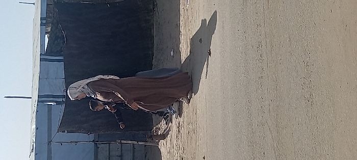 The image depicts a scene in Khan Yunis, Palestinian Territory, under a clear, bright sky. Three individuals are walking from right to left on a dusty, light-colored dirt path that transitions into a paved road in the foreground.
In the mid-ground, a woman walks in the lead, facing left. She wears a light-colored hijab, a long, full-sleeved brown abaya, and light-colored athletic shoes. Slightly behind and to her left, a child, likely male, walks in the same direction, wearing a dark long-sleeved top and dark pants with dark sneakers. The lower legs and feet of a third person, wearing light blue pants and dark shoes, are partially visible behind the woman and child, also walking left. All three cast distinct long shadows on the ground to their left, indicating strong sunlight from the right.
To the left of the path, the ground is uneven and dusty. Behind the individuals, a dark fabric structure, possibly a tarp or tent wall, spans across the mid-ground, creating a dark backdrop. Above and behind this structure, several light-colored temporary shelters or tents, constructed from tarpaulin or corrugated metal sheets, are visible, suggesting a camp environment. A thin, vertical wooden pole or stick projects upwards from behind these structures into the clear blue sky.
