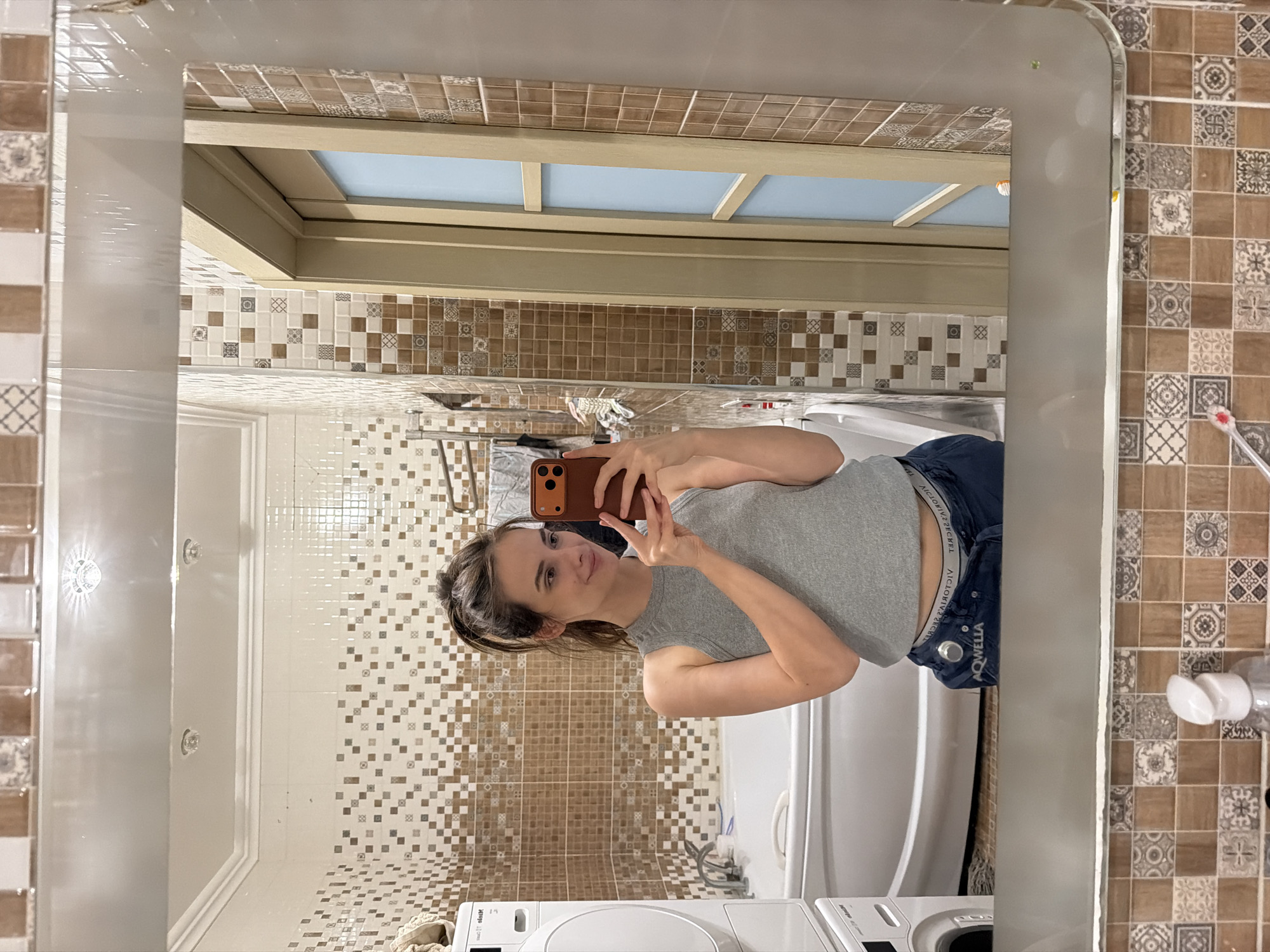This media file is a mirror selfie taken in a bathroom. The primary subject is a young woman with dark hair pulled back, wearing a grey tank top and blue jeans. She is holding a smartphone in her right hand and making a peace sign with her left hand. Her expression is a slight smile.
The bathroom is tiled with a mosaic pattern of brown, beige, and white squares. A white bathtub is visible in the background. To the left of the woman, a white washing machine is partially visible. In the upper part of the reflection, a wooden-framed window with a light blue sky is visible.
The overall impression is casual and personal, suggesting the photo was taken in a home bathroom. There is no indication of the time of day or weather. There is text visible on the woman's underwear band, which appears to read "VICTORIA'S SECRET". Another visible text reads "AQWELLA" on the waistband of her jeans.
