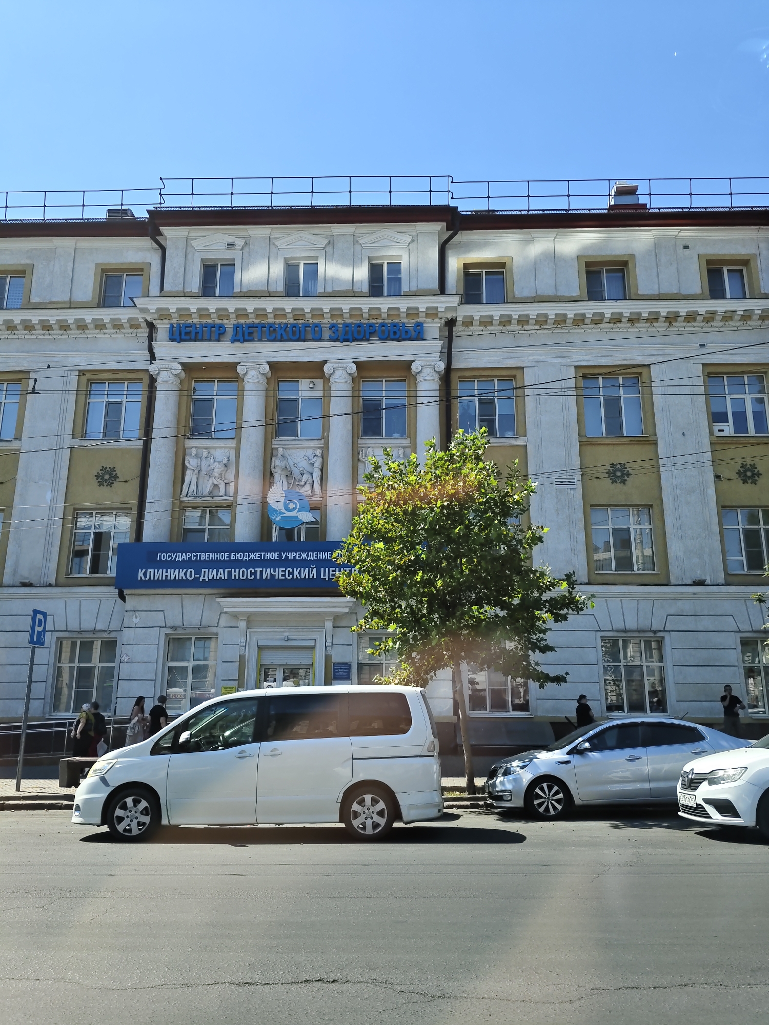 The image depicts the facade of a multi-story building in Rostov-on-Don, Russia, identified by the prominent signage "ЦЕНТР ДЕТСКОГО ЗДОРОВЬЯ" (Center for Children's Health) in blue lettering. Below this, a blue banner reads "ГОСУДАРСТВЕННОЕ БЮДЖЕТНОЕ УЧРЕЖДЕНИЕ" (State Budgetary Institution) and "КЛИНИКО-ДИАГНОСТИЧЕСКИЙ ЦЕНТР" (Clinical Diagnostic Center). The building features classical architectural elements, including columns and decorative friezes. Several windows are visible on its upper floors. In front of the building, a tree with green foliage partially obscures the view. On the street in front of the building, a white minivan is parked, and a silver sedan is also visible, along with a portion of a white car on the far right. Several individuals are seen on the sidewalk, including a woman with a bag, and a man in black. The sky is clear and blue, indicating daytime. The scene is captured from a street-level perspective, likely from another vehicle given the angle.