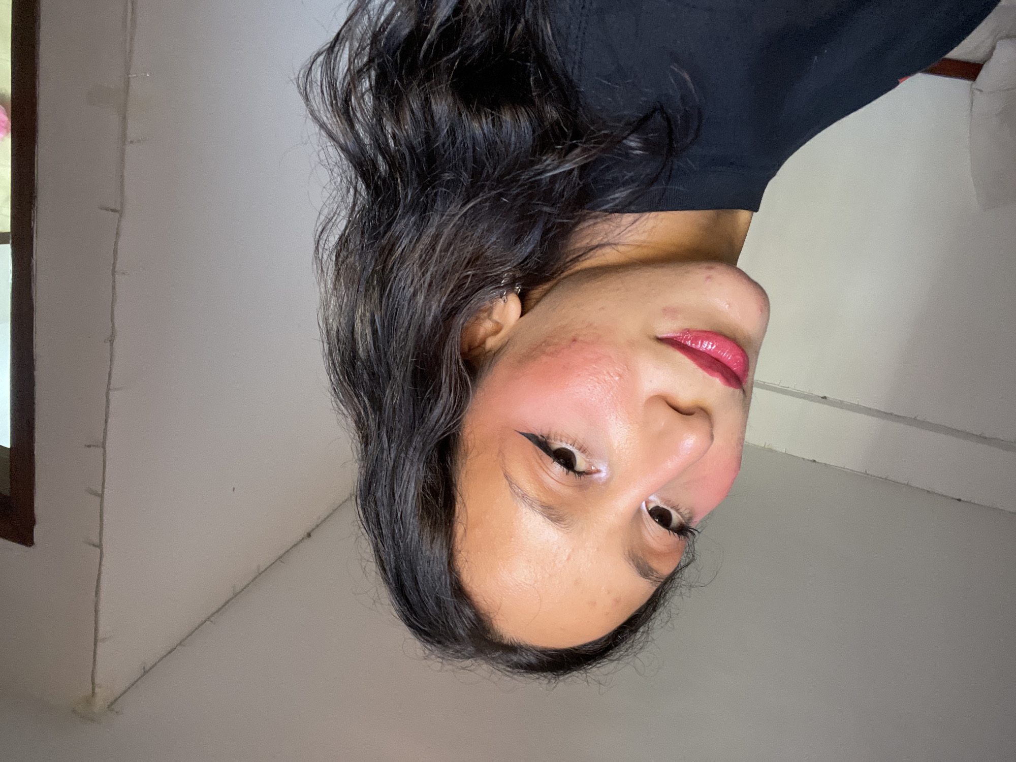 A young woman with dark, wavy hair and light skin is posing for a selfie. She is wearing a black top and has on makeup including pink blush, eyeliner, and glossy pink lipstick. She has a light-colored wall and what appears to be a window with wooden trim behind her. There are strings of small lights on the wall. The lighting suggests she is indoors, possibly in a home or hotel room in Porlamar, Venezuela.