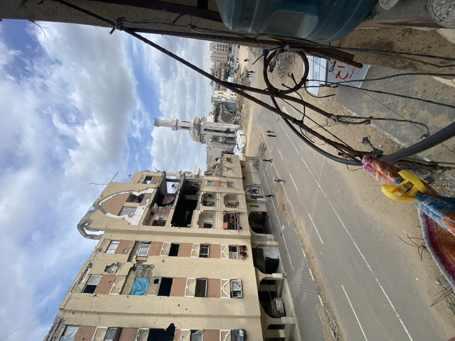 The image depicts a street view in Khan Yunis, Palestinian Territory, showing widespread structural damage under a partly cloudy sky. In the foreground, from an elevated perspective, a dark metal balcony railing with intricate scrollwork and tangled black wires is visible on the right. A light blue plastic water jug is partially seen on the far right, and a multi-colored cloth with a yellow clothespin hangs from the railing at the bottom.
The mid-ground features a two-lane asphalt road, partially covered in dirt on its shoulders, where several individuals are walking. To the left of the road, a multi-story residential building, light brown with peach-colored decorative elements, exhibits extensive damage. Numerous bullet holes or shrapnel impacts mar its facade, and large sections of walls and windows are destroyed or missing, exposing interior rooms. A blue tarp hangs from one upper-level opening, and a silver sedan is parked beneath a ground-floor archway.
In the background, dominating the center, stands a severely damaged mosque. Its minaret, cylindrical and light-colored, remains upright but shows significant structural compromise. The main mosque structure is largely reduced to rubble, with its dome and walls extensively collapsed, surrounded by large piles of debris. Multiple individuals are visible walking on the cleared ground around the mosque. Further in the distance, additional urban structures, some appearing damaged, complete the horizon.
