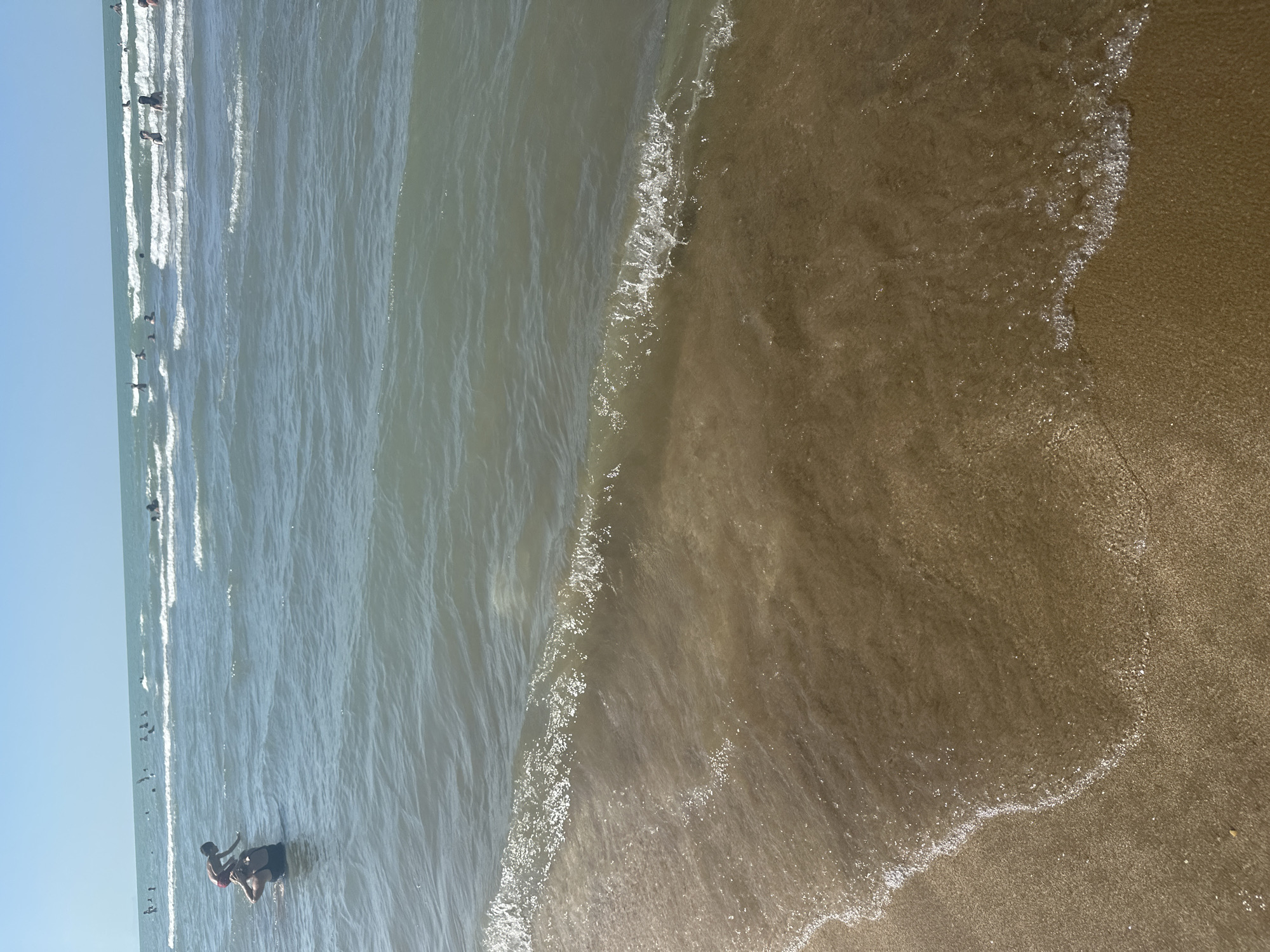 A wide-angle shot captures a sunny day at a beach, with the ocean dominating the frame. The water is a murky green-blue, with small, choppy waves rolling towards a sandy shore. The sand in the foreground is wet and brown, marked by the receding tide.
In the middle ground, several people are visible in the water. To the left, an adult in a dark swimsuit appears to be carrying a child on their shoulders. The child is wearing red shorts. Further out in the water, other individuals are swimming and playing in the waves. The sky is a clear, bright blue.
The scene suggests a casual day at the beach, with people enjoying the water under a bright sky. The location is identified as Sabunchu Raion, Azerbaijan, a district known for its coastal areas along the Caspian Sea.