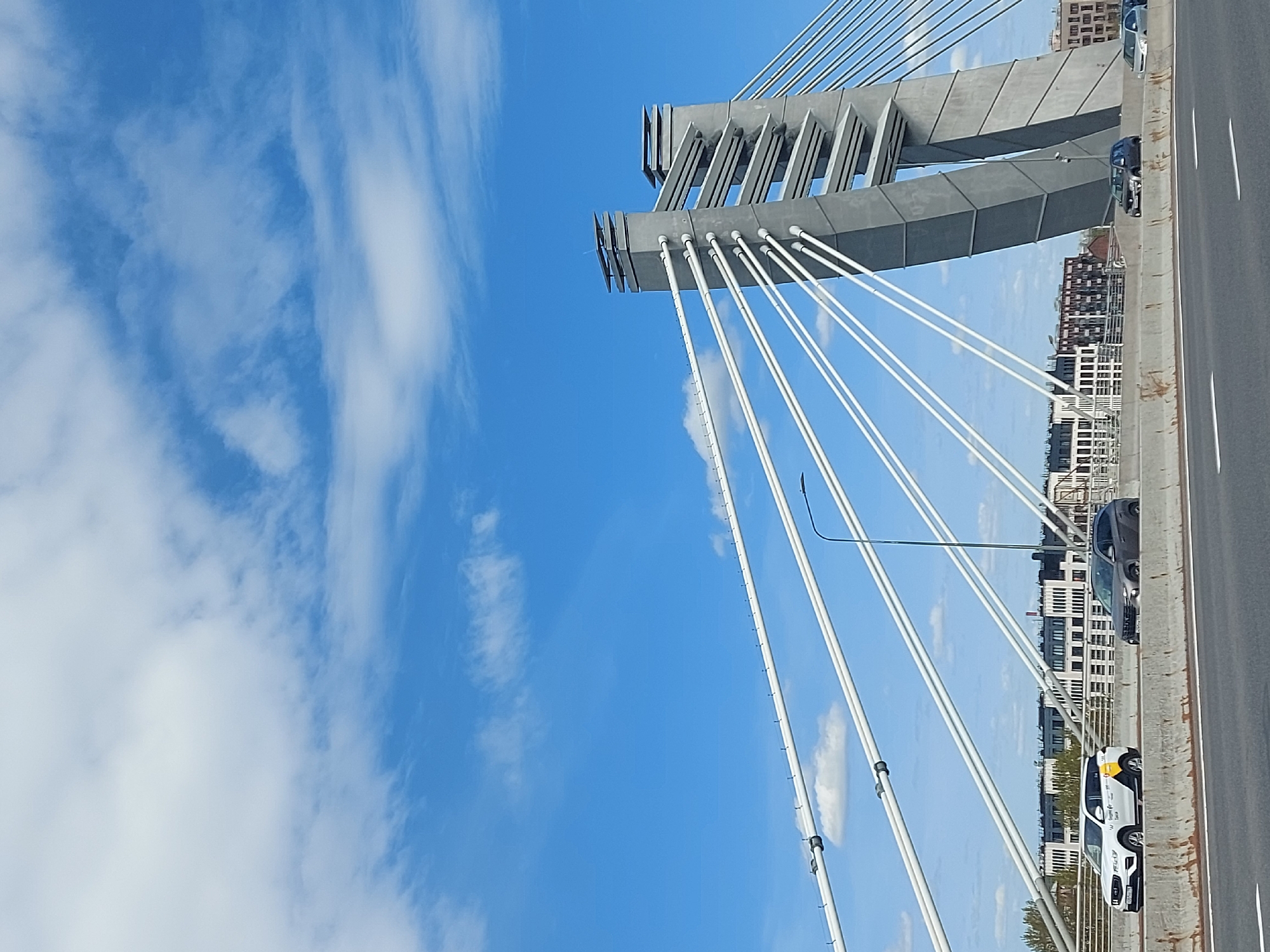 The image depicts a section of a cable-stayed bridge in Saint Petersburg, Russia, possibly the Western High-Speed Diameter (WHSD) bridge. The view is angled upwards toward the bridge's support structure and cables against a blue sky with scattered clouds. The bridge's roadway is visible on the right, with several cars traveling on it. Buildings of the city are visible in the background, beyond the edge of the roadway. The bridge appears to be under normal vehicular traffic.