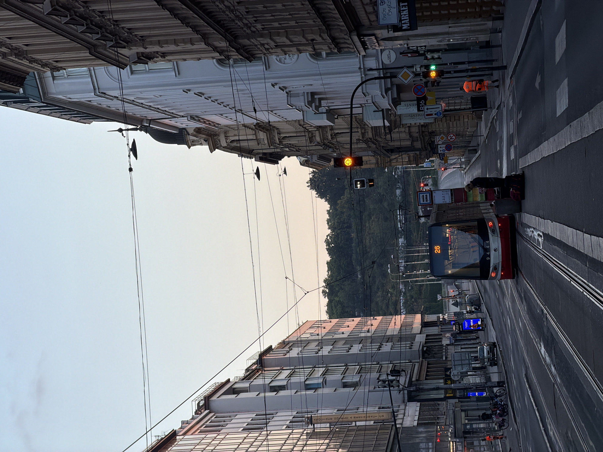 The image depicts a street scene in Prague, Czechia, likely during the late afternoon or early evening. The perspective is from the ground, looking down a street lined with buildings on both sides. The street is a tram track with a red and white tram numbered "26" approaching the viewer. The tram is in the center of the frame, on the right-hand set of tracks.
On the left side of the street, there is a multi-story building with a glass facade reflecting the light. Further down, another building with many windows is visible. There are several parked bicycles and a car on the left side of the street. A traffic light is present near the intersection.
On the right side of the street, there are older, ornate buildings with architectural details. A traffic light is illuminated with a yellow arrow pointing upwards. Several street signs are visible, including one with "R16" and a symbol indicating a prohibition. A person wearing an orange high-visibility vest is standing on the sidewalk. A sign with "MARKET" is visible on a storefront.
Two people are standing on the tram tracks near the approaching tram, one with luggage. A large, dark hill covered in trees is visible in the background, beyond the buildings. The sky is a pale blue with a hint of pink at the horizon, suggesting either sunrise or sunset. Overhead, numerous power lines crisscross the scene. The overall impression is a busy urban environment with ongoing public transportation.